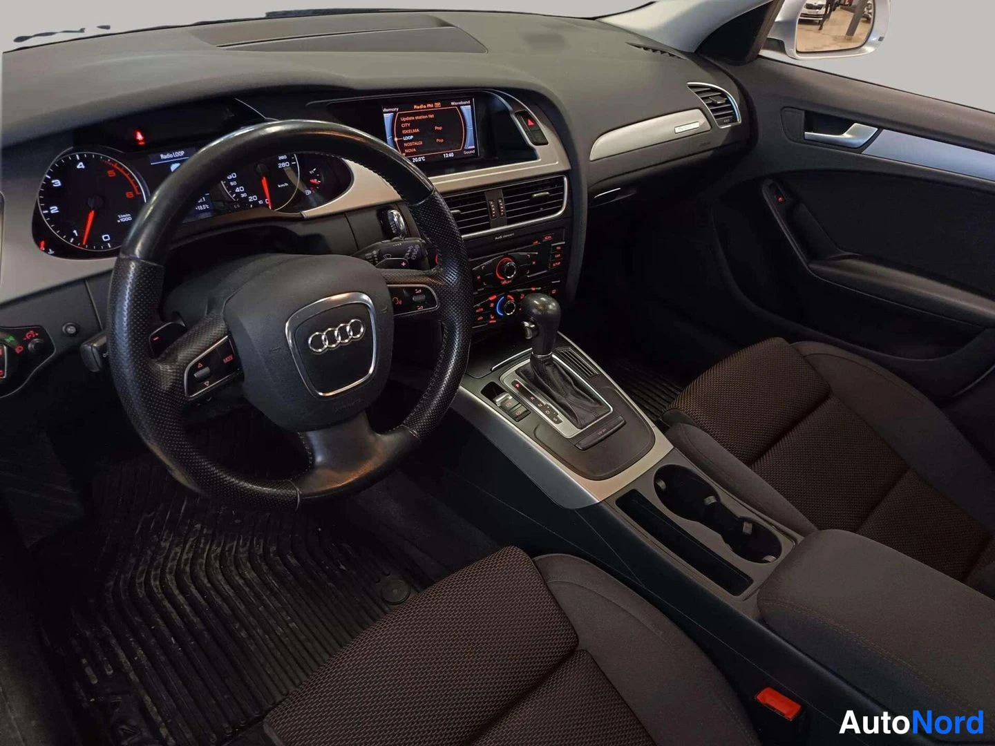 Edestä: Auton ohjaamo: musta nahkainen ohjauspyörä Audi-renkailla, mittaristo, keskiteline automaattivalitsimella, infotainment-näyttö ja tummat kangasistuimet.
