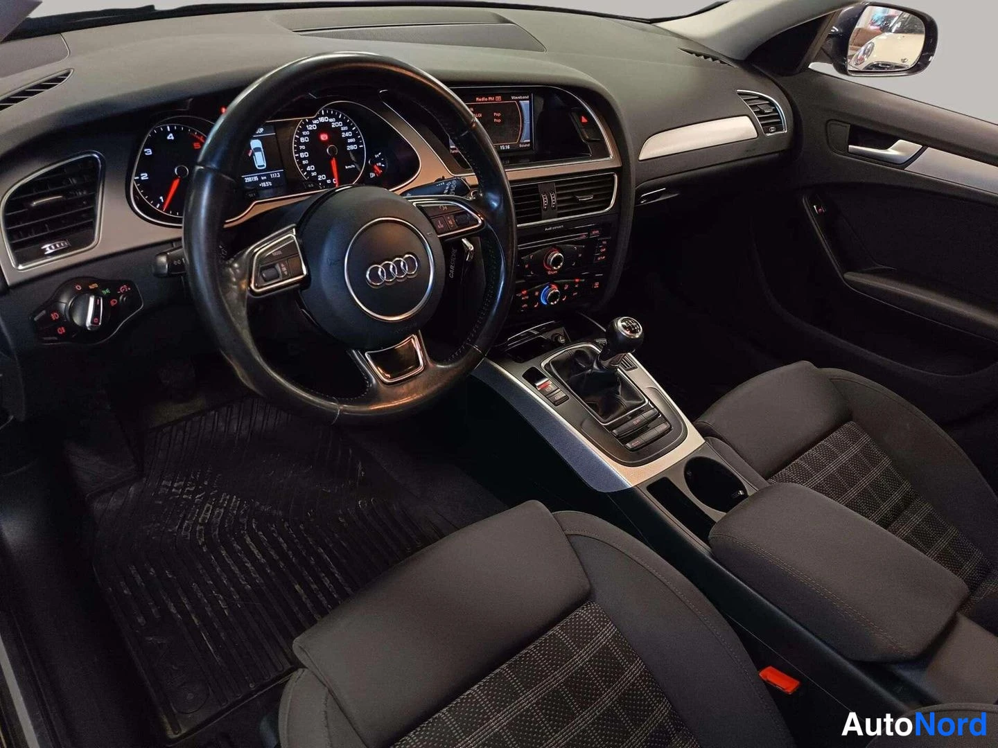 Muu: Auton sisätilat kuljettajan puolelta: Audi-ohjauspyörä, mittaristo, keskikonsoli manuaalivaihteistolla ja tummat kangasistuimet.