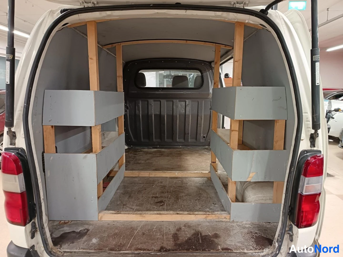 Tavaratila: Rear view of an empty cargo area inside a white van with tailgate open. The space is fitted with grey shelving units on both sides and wooden framing supports.