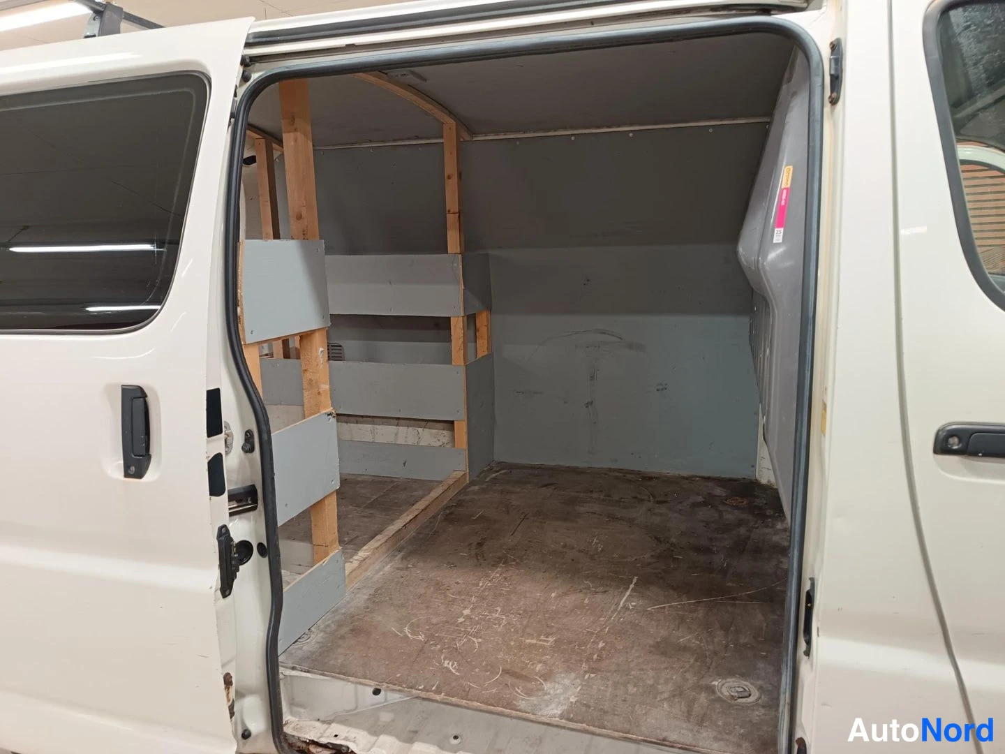 Tavaratila: Side view of open cargo doors revealing an empty white van interior with grey shelves and wooden framing supports along the left side. Floor shows wear marks. C