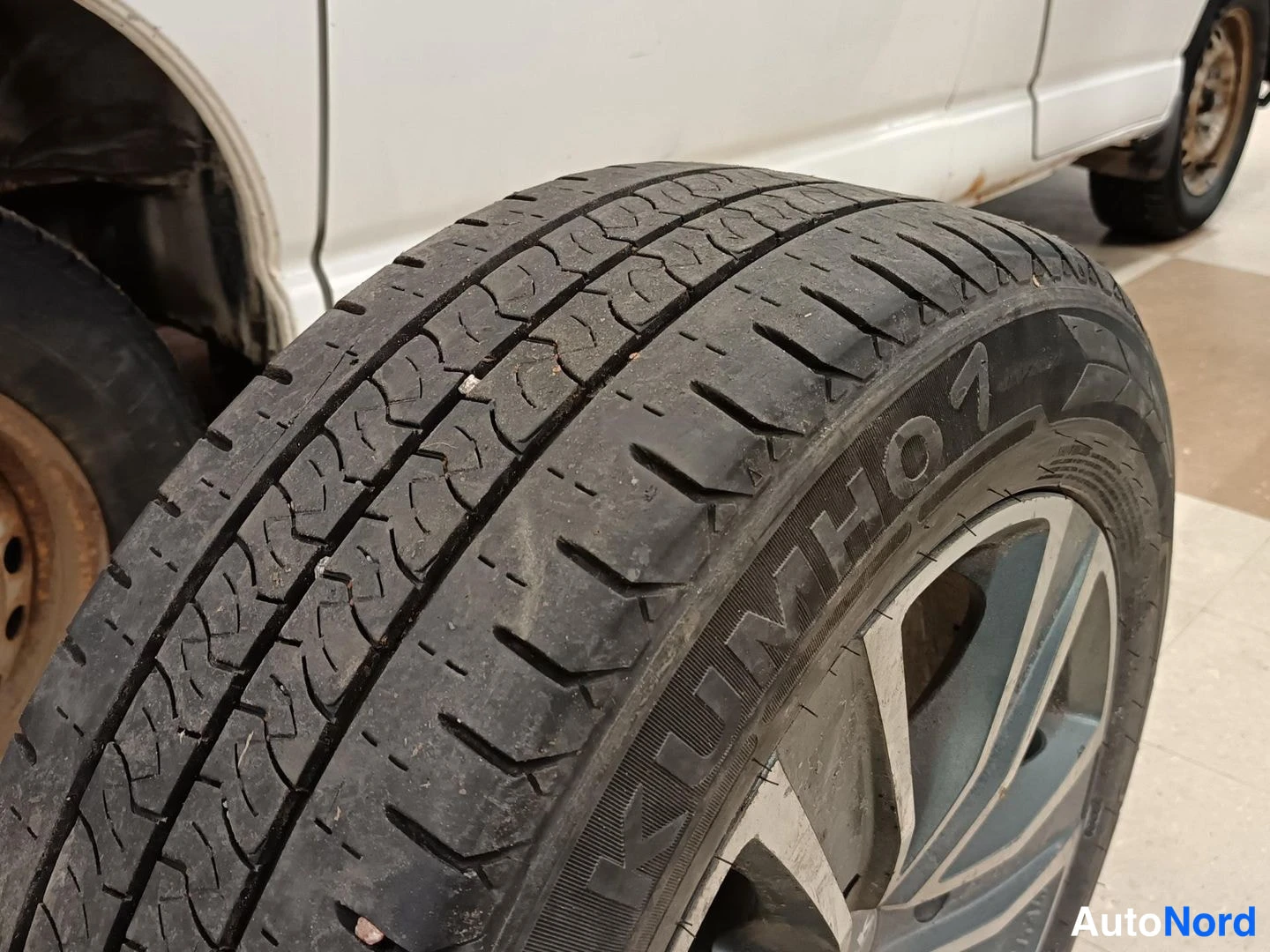 Muu: Lähikuva Kumho-renkaasta, vieressä auton valkoinen koripaneeli ja ruostunut vanteen reuna.