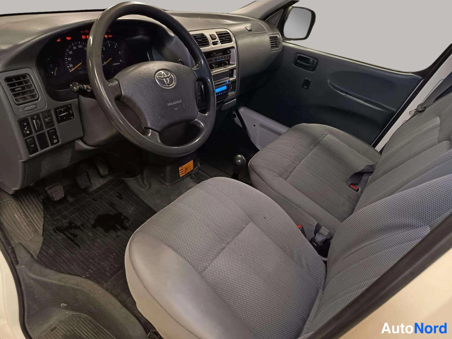 Muu: Auton ohjaamo kuljettajan puolelta: Toyota-ohjauspyörä, mittaristo ja harmaat kankaaverhoillut istuimet.