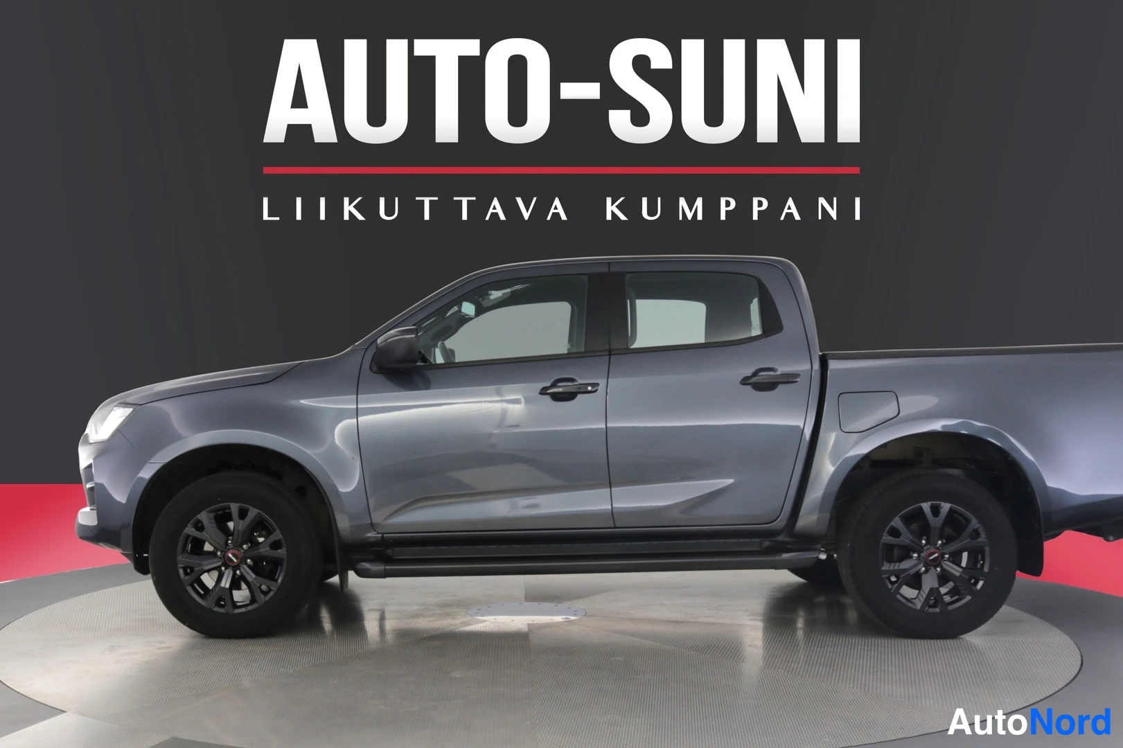 Sivulta: Tummanharmaa Isuzu D-Max -avolava-auto kuvattuna vasemmalta etukulmasta showroomin pyöräalustalla; mustat kevytmetallivanteet, astinlaudat ja tummat la