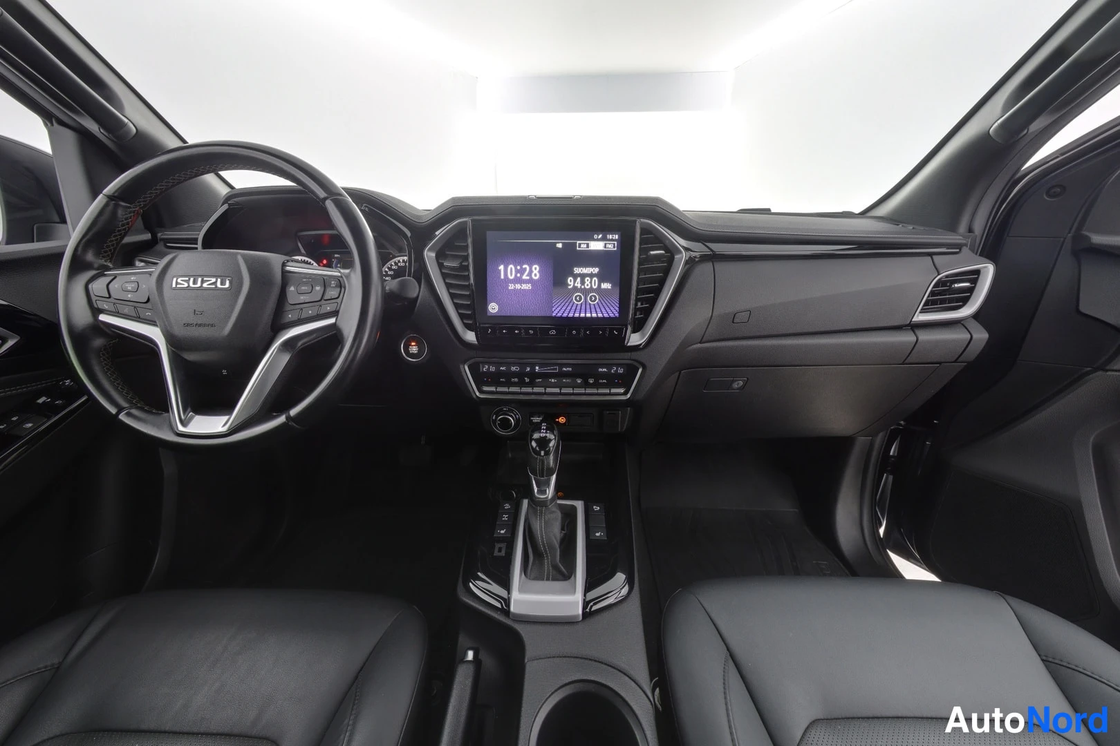 Sivulta: Auton sisätilat edestä: Isuzu-merkinen ohjauspyörä, kojelauta, infotainment-näyttö, automaattivaihteiston valitsin ja mustat istuimet.
