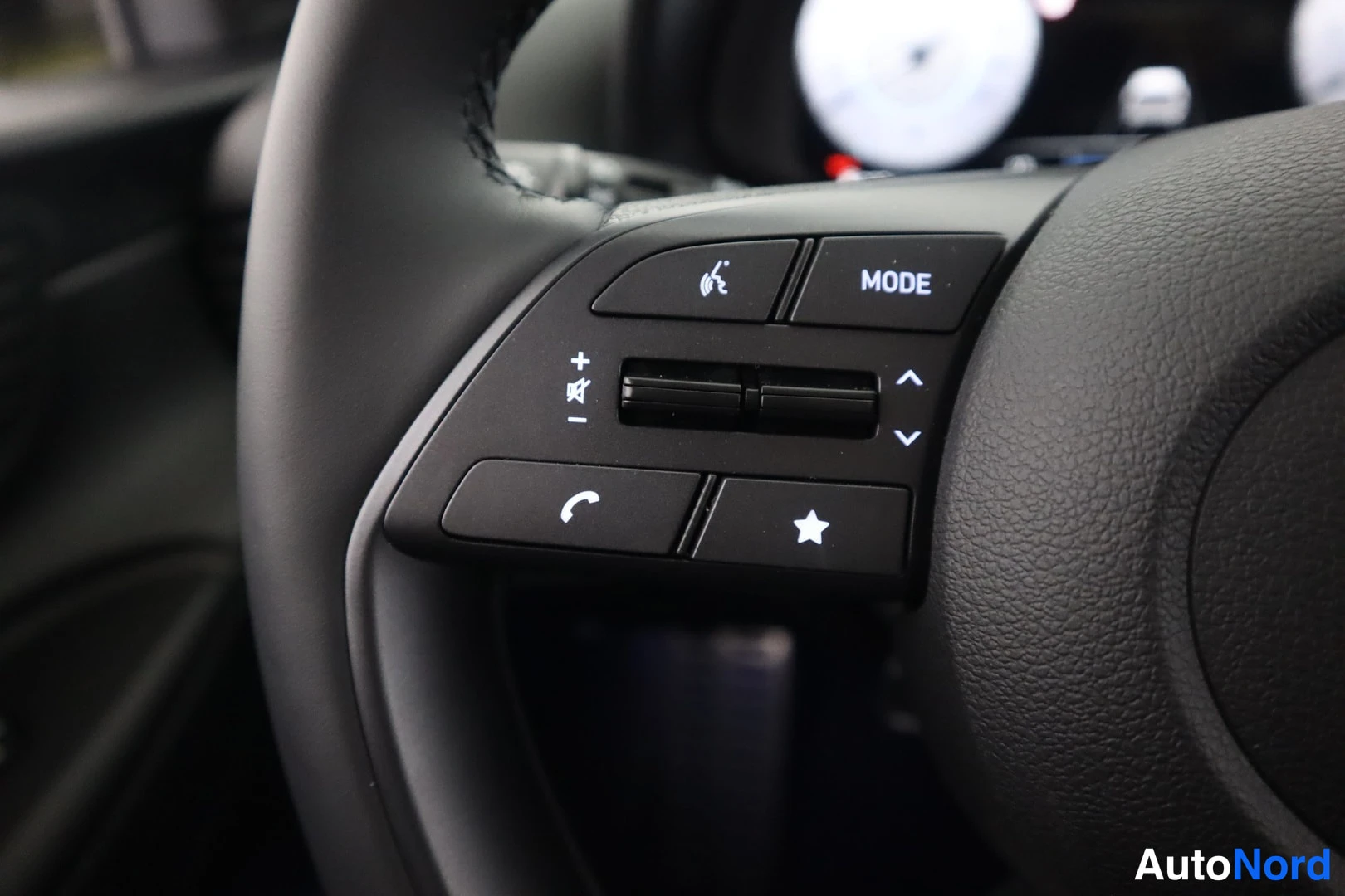 Yksityiskohta: Detailed view of the left side of the steering wheel showing buttons for voice control, audio modes (MODE), volume adjustment (+/-) and phone call management.