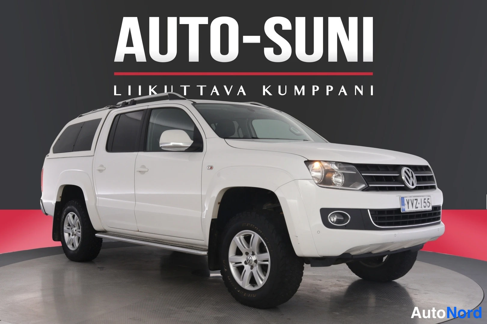 Etuviisto, nokka oikealle: Valkoinen Volkswagen Amarok-neliovinen pickup etukulmasta oikealta, roofbox ei, takalavalla kannellinen kovakuori ja kattotelineet.