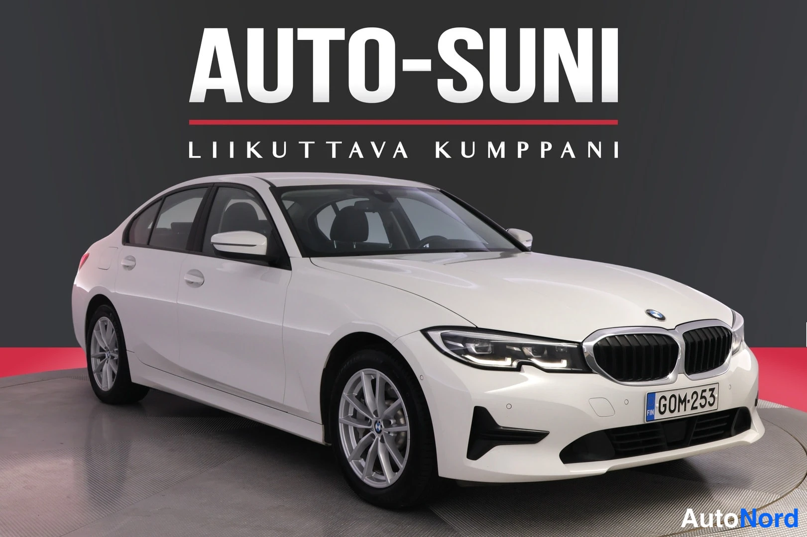 Etuviisto, nokka oikealle: valkoinen sedan kuvattuna etukulmasta, rekisterikilpi, taustalla AUTO-SUNI-logo