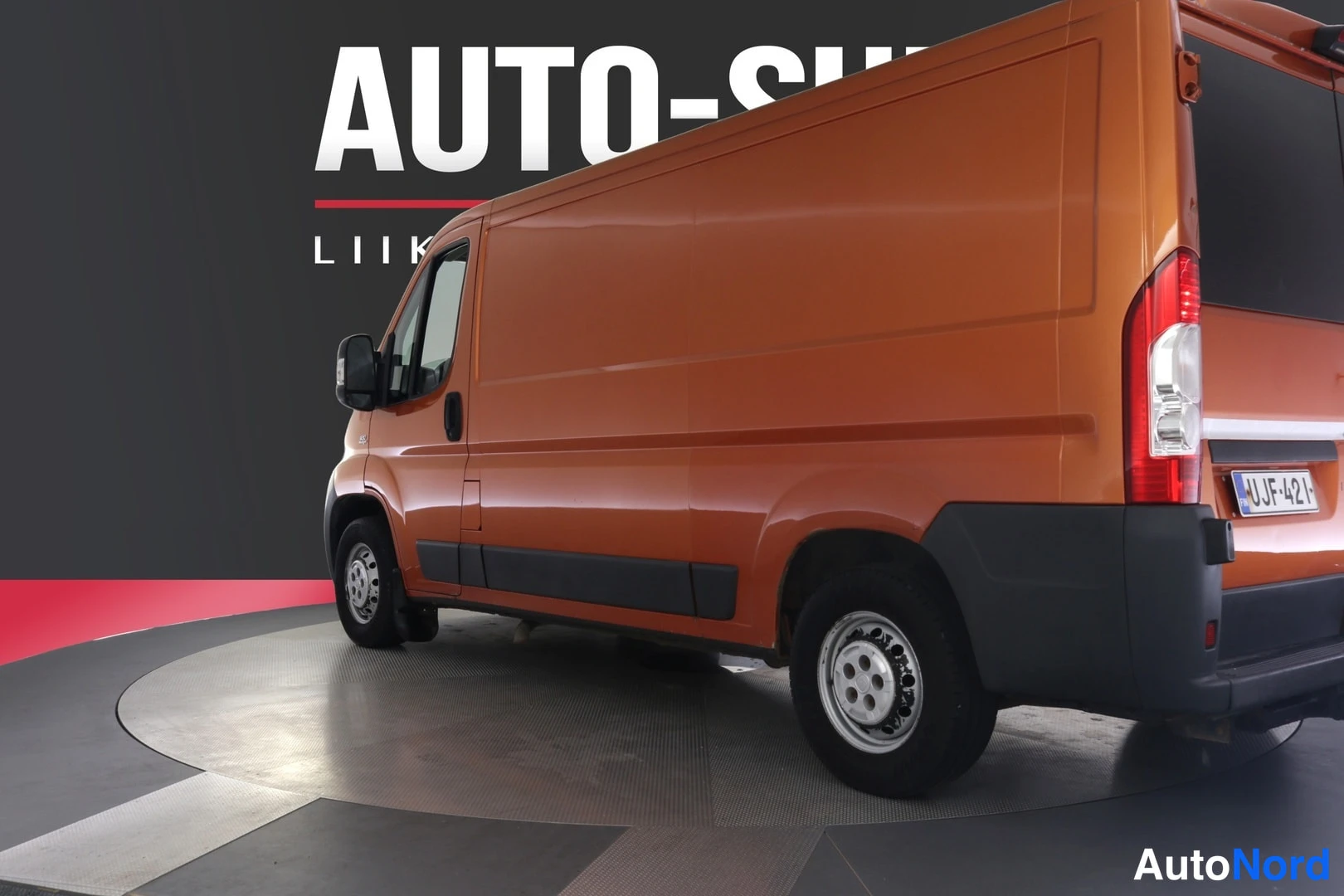 Takaviisto: Oranssi Fiat Ducato -pakettiauto takakulmasta oikealta kuvattuna studiossa, -kilpi näkyvissä.