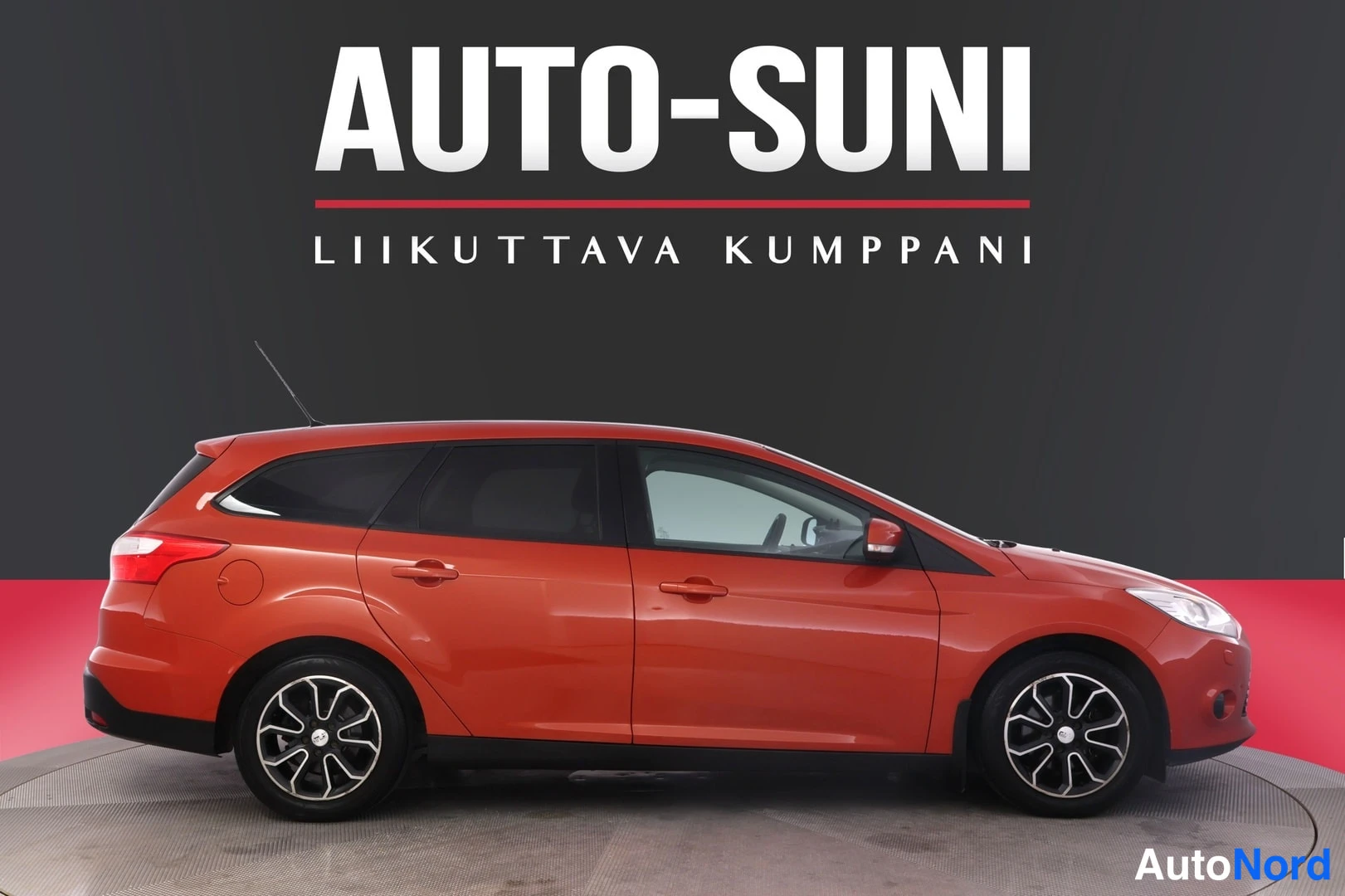 Etuviisto, nokka oikealle: Oranssinpunainen Ford Focus -farmari sivulta etukulmaan, keulakko oikealla, tummassa studioympäristössä Auto-Suni-mainostekstillä.