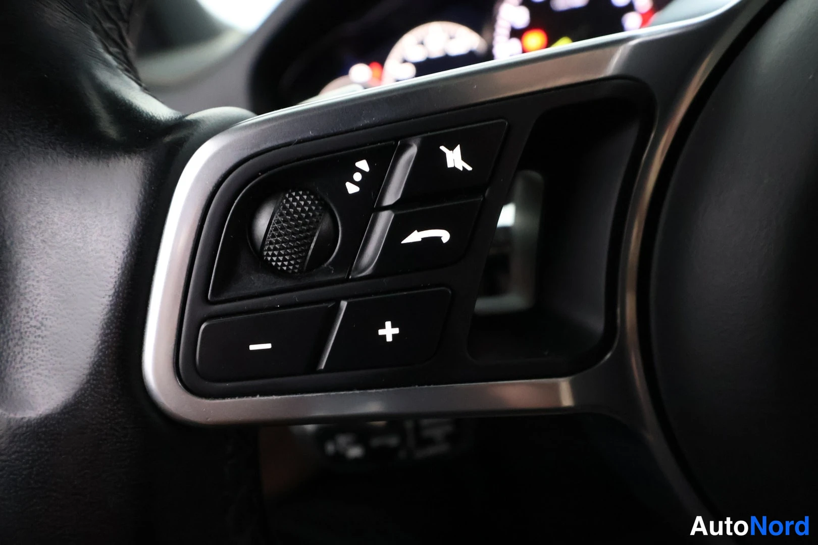 Yksityiskohta: Close-up of the right-hand spoke of a leather-wrapped steering wheel showing multimedia controls including a scroll wheel, volume +/- buttons, mute, and call an