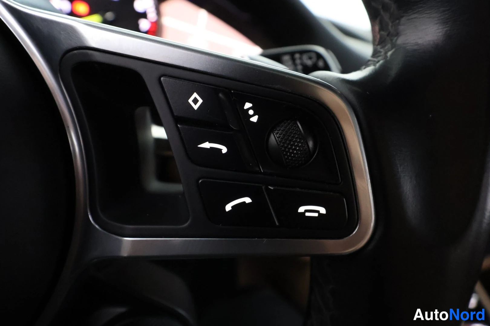 Yksityiskohta: Close-up of the left-hand spoke of a black leather-wrapped steering wheel showing menu navigation buttons, dial, and phone hang-up/receive icons.