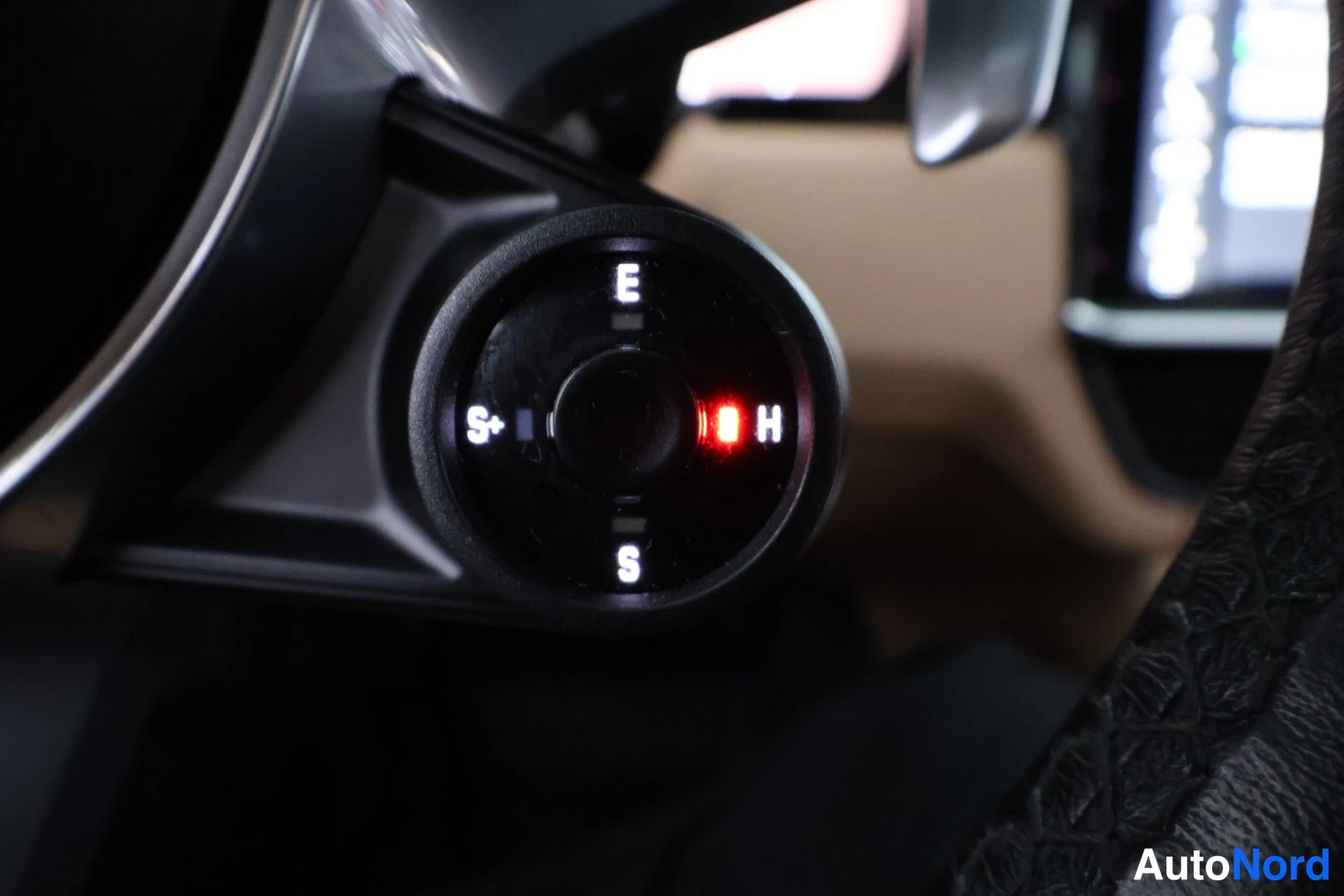 Yksityiskohta: Circular drive mode selector knob on the center console illuminated in red, with markings for Economy (E), Sport (S), Sport+ (S+) and Hand (H).
