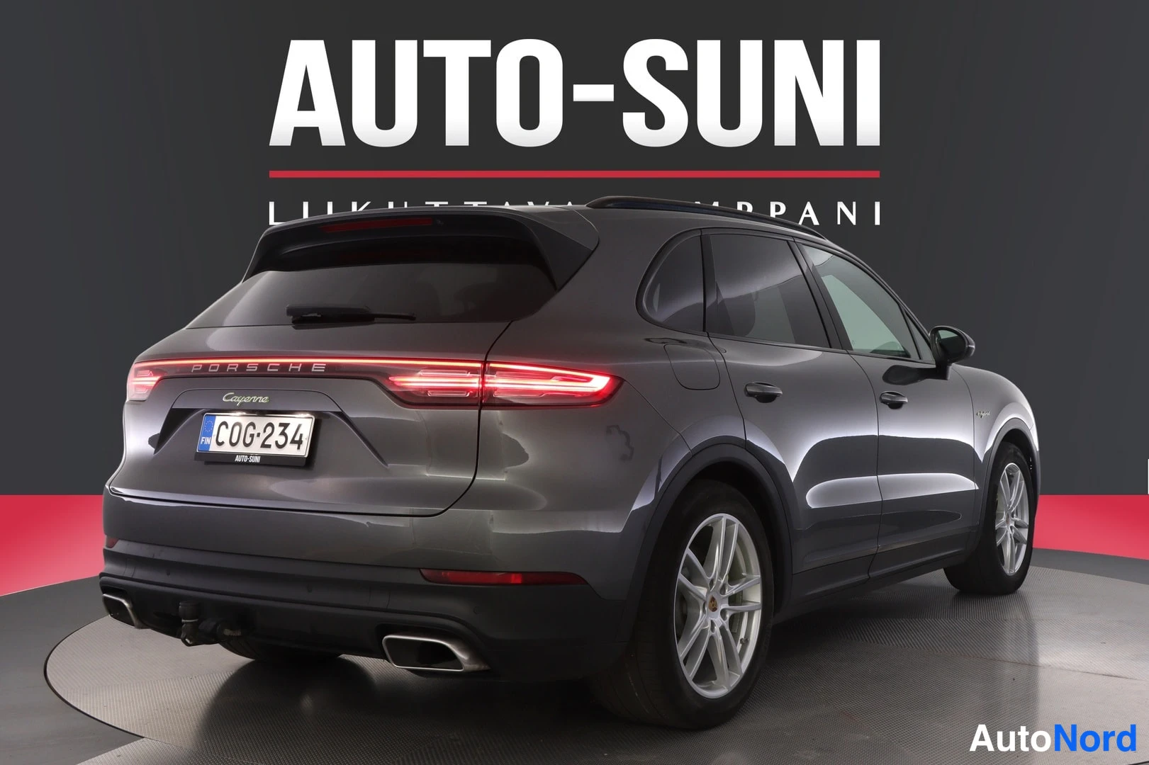 Takaviisto: Porsche Cayenne 2020, tummanharmaa auto takaa oikealta kuvattuna, rekisteritunnus, takavalo syttyneenä, taustana AUTO-SUNI-mainos.