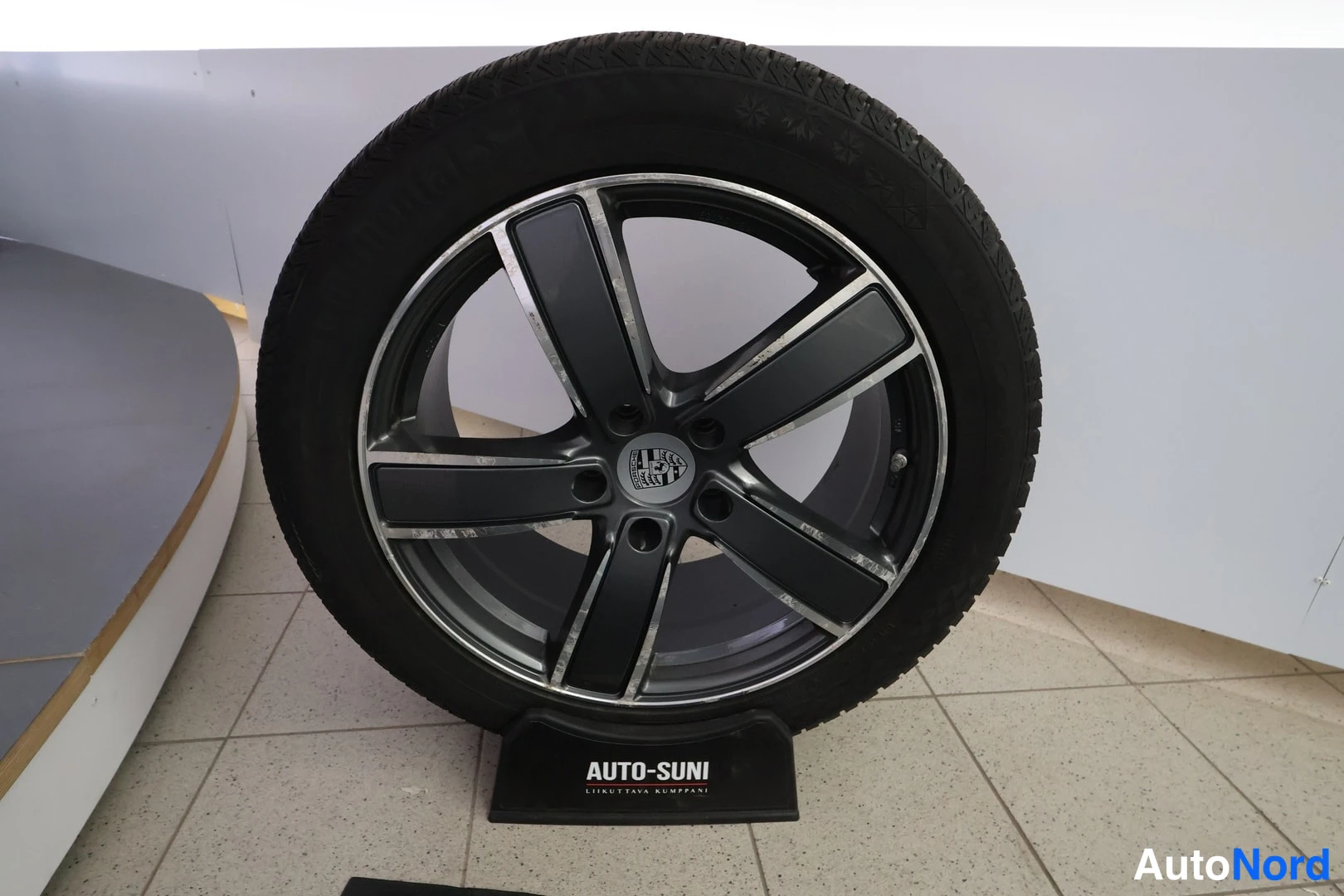 Muu: Musta alumiinivanne viidellä parralla Porsche-emblemillä keskiössä, talvirenkaalla, näytöllä AUTO-SUNI-telineessä.