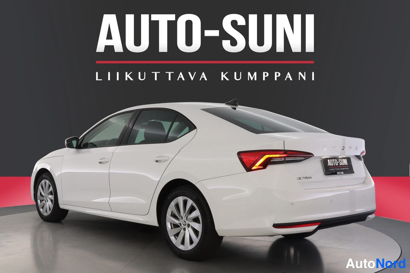 Takaviisto: Valkoinen sedan-henkilöauto takaosalta ja vasemmalta sivulta neljästä kolmasosaan takakulmasta, perä oikealla, showroom-taustassa.