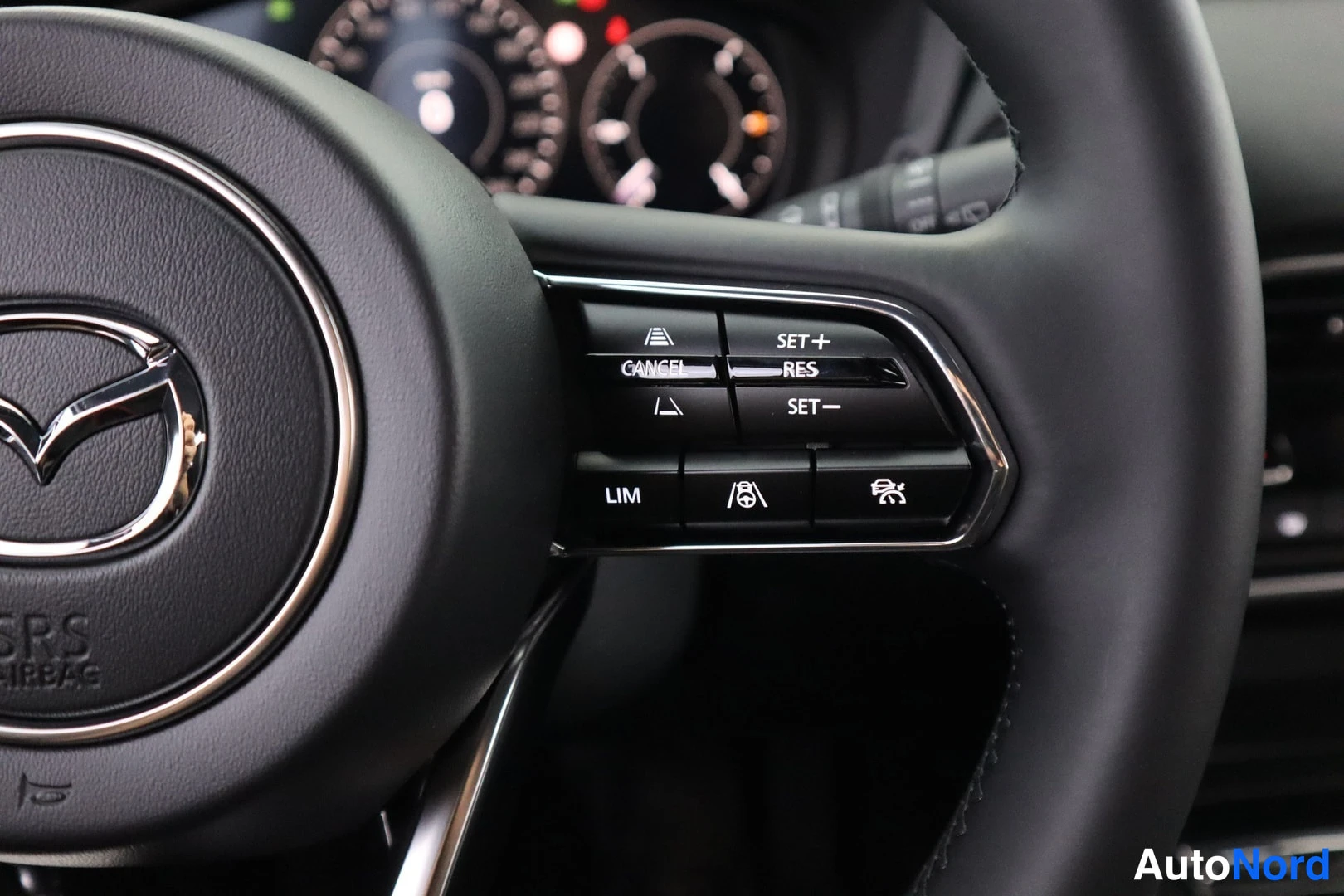 Muu: Mazda-ohjauspyörän vasen puhelin: nappirivi teksteillä SET+, RES, SET−, CANCEL ja LIM, taustalla mittaristo.