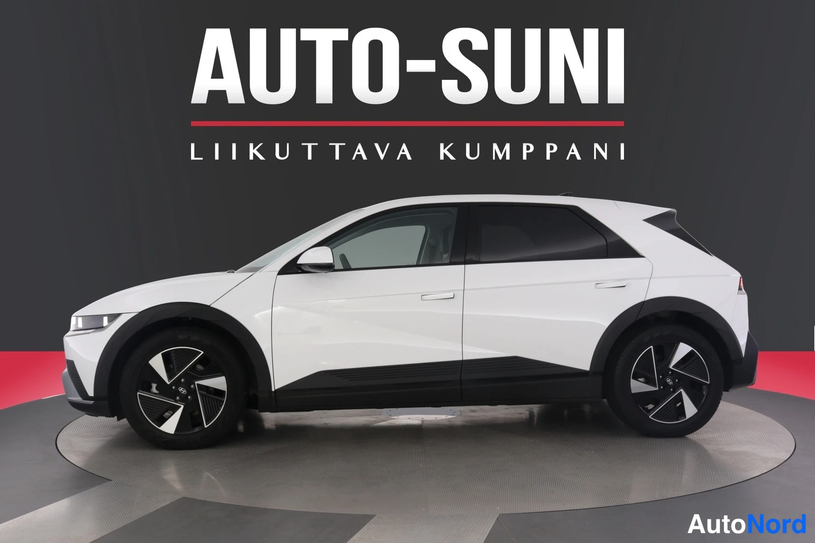Sivulta: Sivukuva valkoisesta Hyundai IONIQ 5 -sähköautoa esittelevässä studioympäristössä, Auto-Sunin taustaseinän edessä.