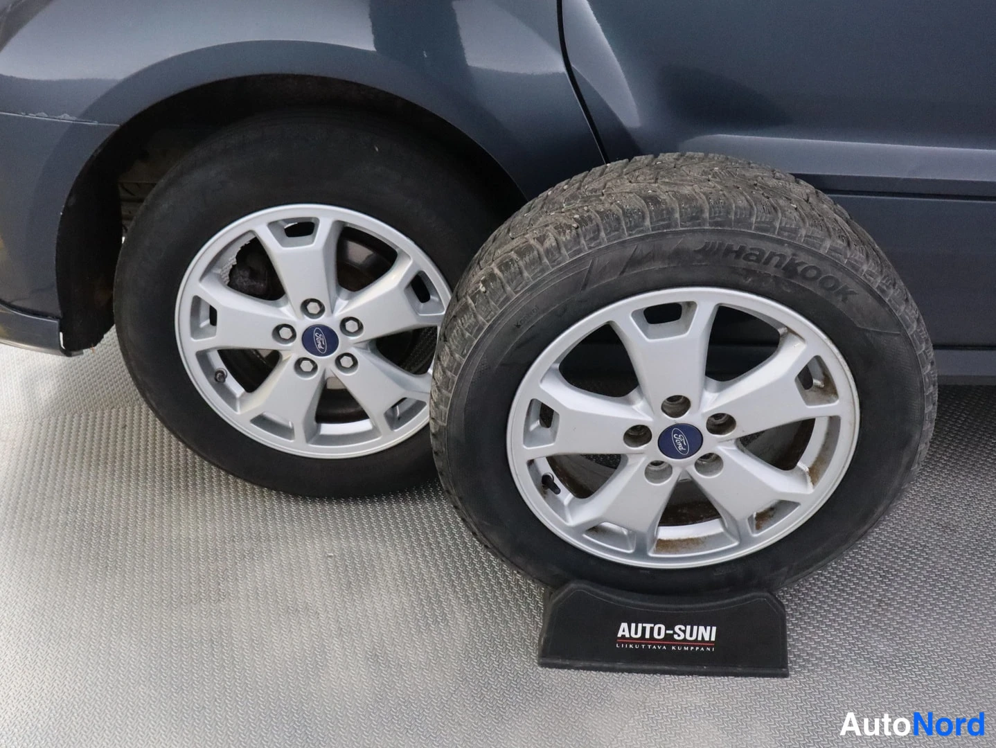 Yksityiskohta: Lähikuva autossa kiinnitetystä hopeasta viisipolttovanteesta Ford-keskiömerkillä sekä vieressä maassa olevasta Hankook-nastarenkaasta samanlaisella vanteella.