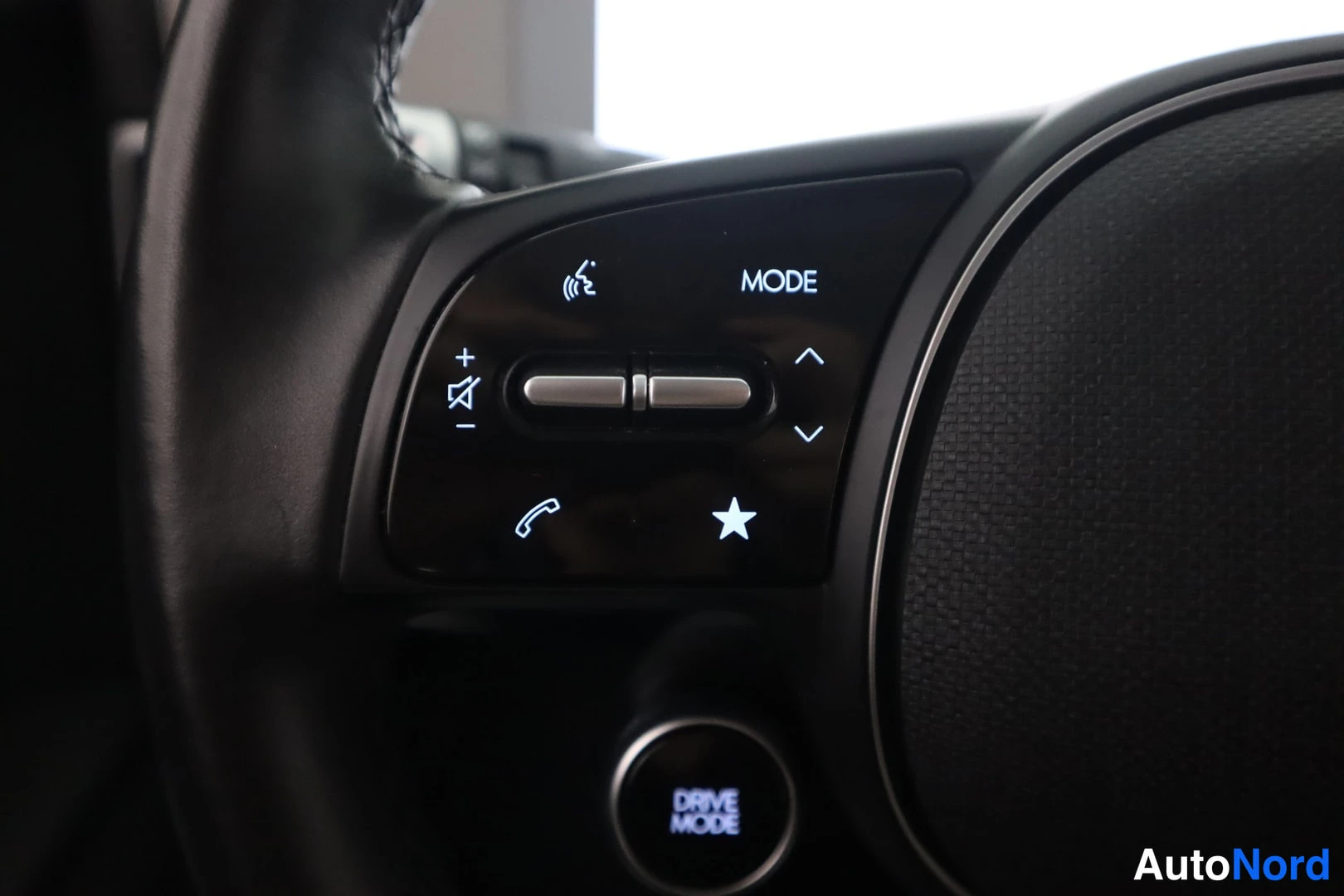 Muu: Ohjaustestin vasemman puolen näppäinryhmä lähikuvassa, MODE-teksti ja puhelin- ja tähtikuvakkeet valaistuna, alapuolella DRIVE MODE -pyöritsäädin.
