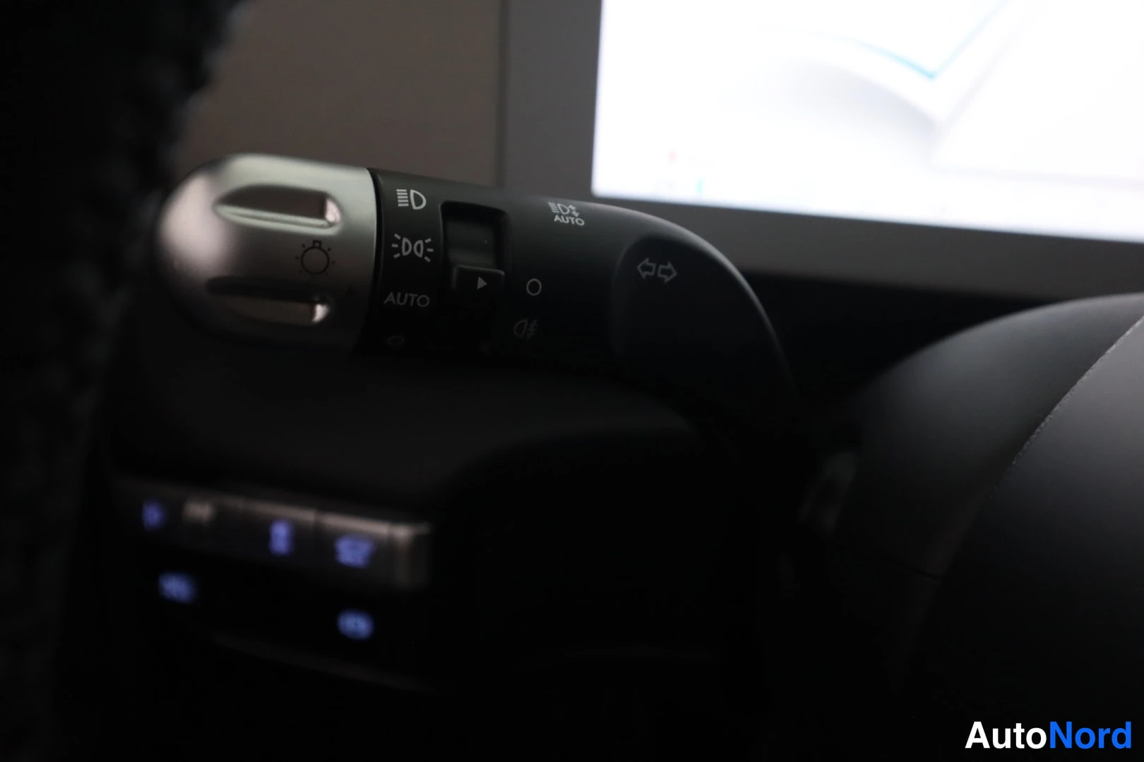 Muu: Vakosamuttimen ja valokytkimen varsilähikuva pimeässä ohjaamossa, AUTO-merkintä ja AUTO HIGH BEAM -symboli, taustalla sumea näyttö.