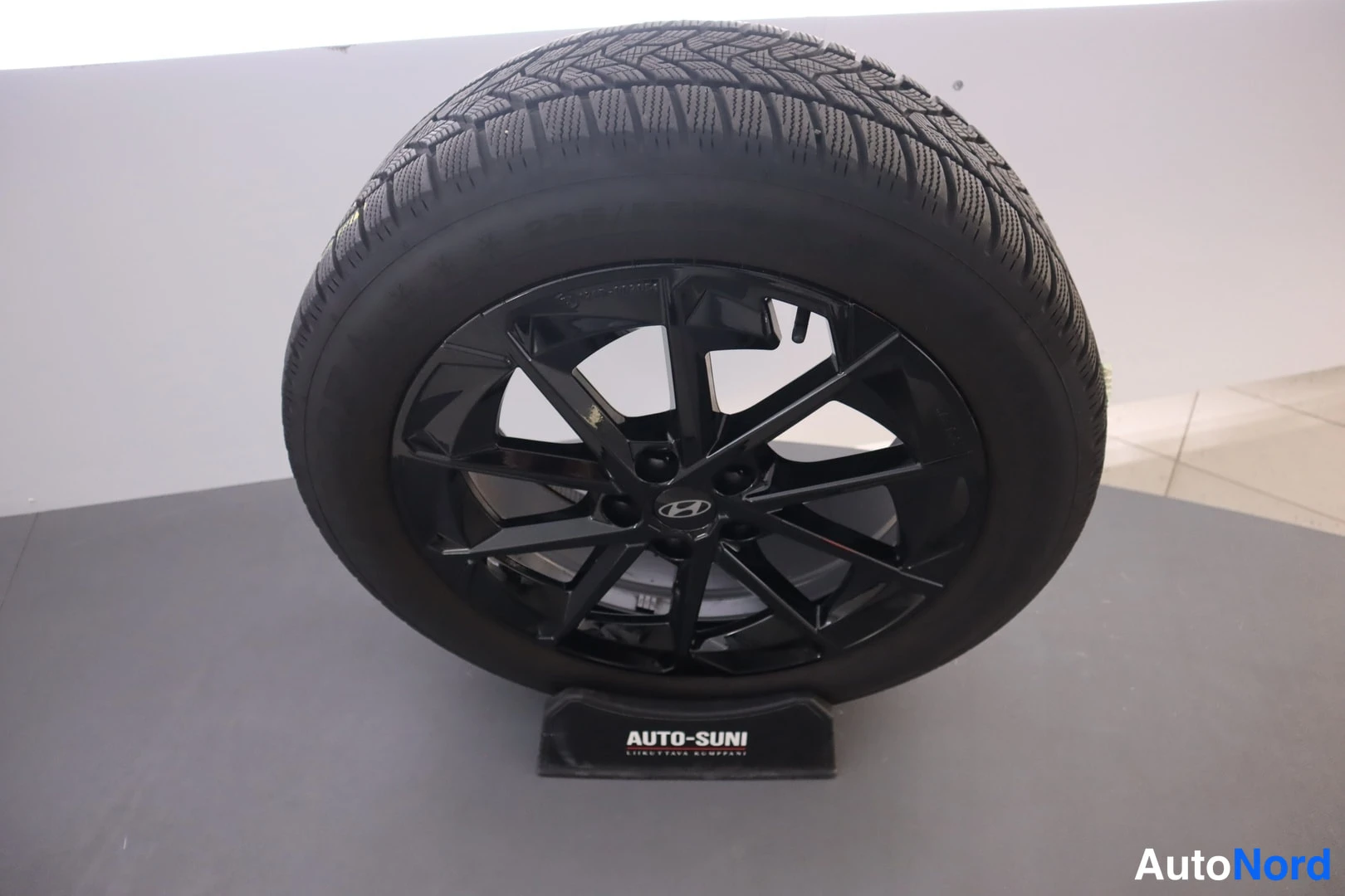 Muu: Hyundai-merkkinen musta kelkkavanteinen talvirengas 225/55 R 19 näytteistelineessä AUTOSUNI-kaupan lattialla.