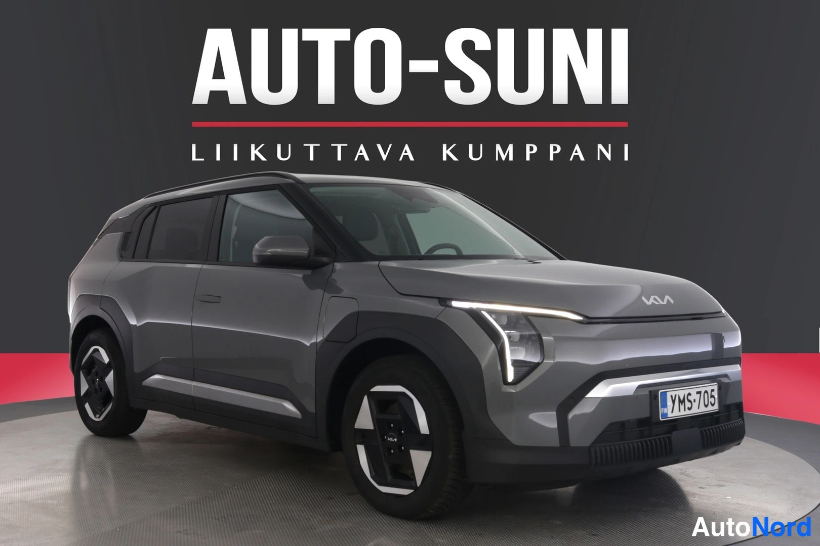 Etuviisto, nokka oikealle: Auton etukulma valokuvausstudiossa, harmaanruskea Kia EV3, rekisteritunnus, AUTO-SUNI-logo taustalla.