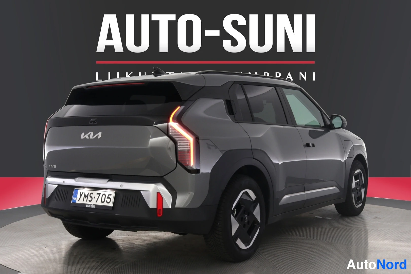 Takaviisto: Auton takakulma valokuvausstudiossa, harmaanruskean Kia EV3:n takavalot ja rekisteritunnus, AUTO-SUNI-logo taustalla.