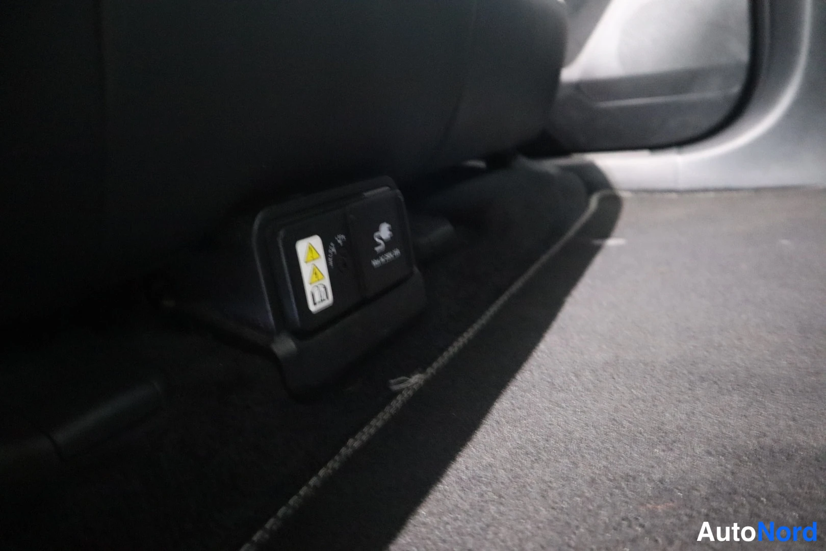 Yksityiskohta: Musta istuinperä ja lattiamatto, jonka vieressä näkyvät lastenistuimen kiinnikkeiden (ISOFIX) merkinnät ja vaihdelaatikon/nappipaneeli tummassa sisätilassa.