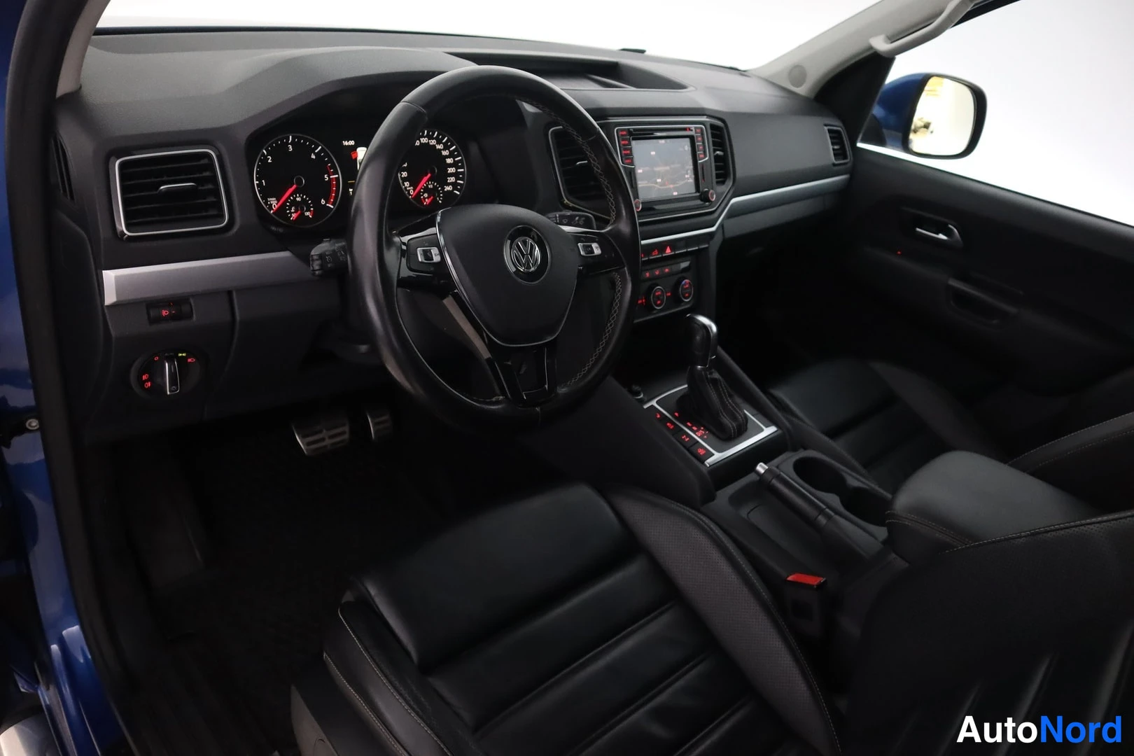 Edestä: Auton etuohjaamo: Volkswagen-merkkinen ohjauspyörä, analoginen mittaristo ja keskikonsolin infotainment-näyttö. Mustat istuimet, automaattivaihteen valitsin ja