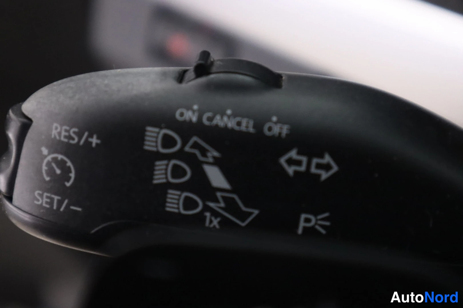 Yksityiskohta: Close-up photograph of a black plastic multi-function stalk on the steering column showing white printed labels 'RES/+' and 'SET/-' for cruise control, symbols