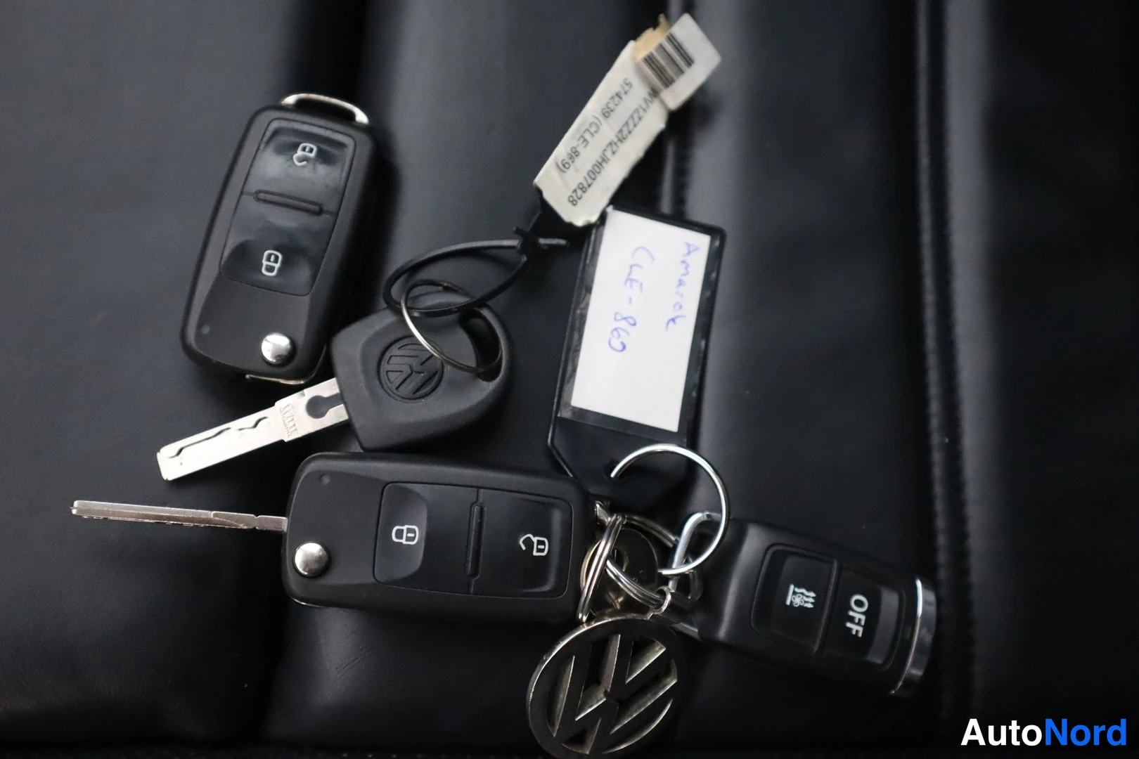 Yksityiskohta: Mustat auton avaimet ja kaksi kauko-ohjainta avainnipussa VW-logon kera, taustalla mustaa nahkaa ja paperilappu, jossa käsinkirjoitettu teksti.