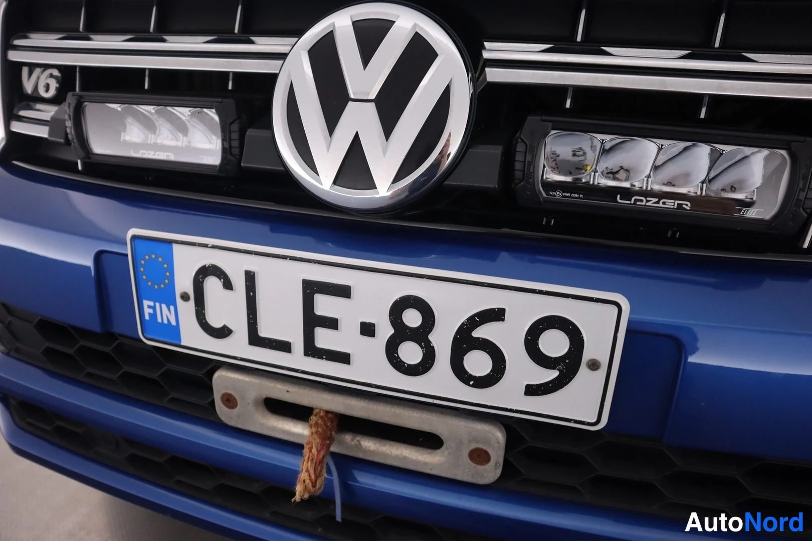 Edestä: Sinisen auton etuosan lähi kuva: VW-merkki, V6-tunnus, kaksi Lazer-tyyppistä lisävaloa ja rekisterikilpi, alla hinauskoukku köysineen.