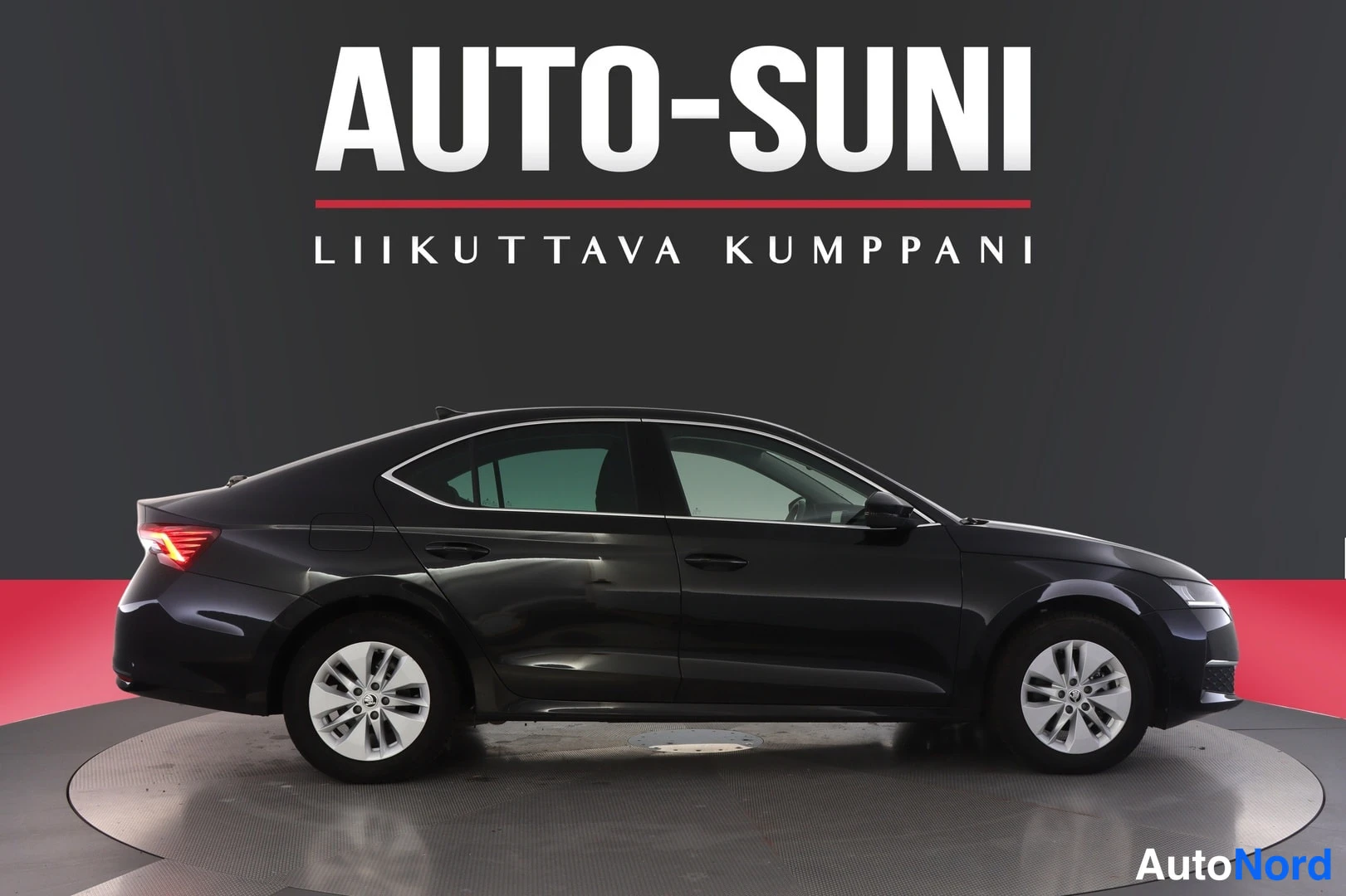 Sivulta: Musta sedan sivusta nähtynä kiertopöydällä sisätiloissa. Kuvassa on myös taustalla AUTO-SUNI -teksti.