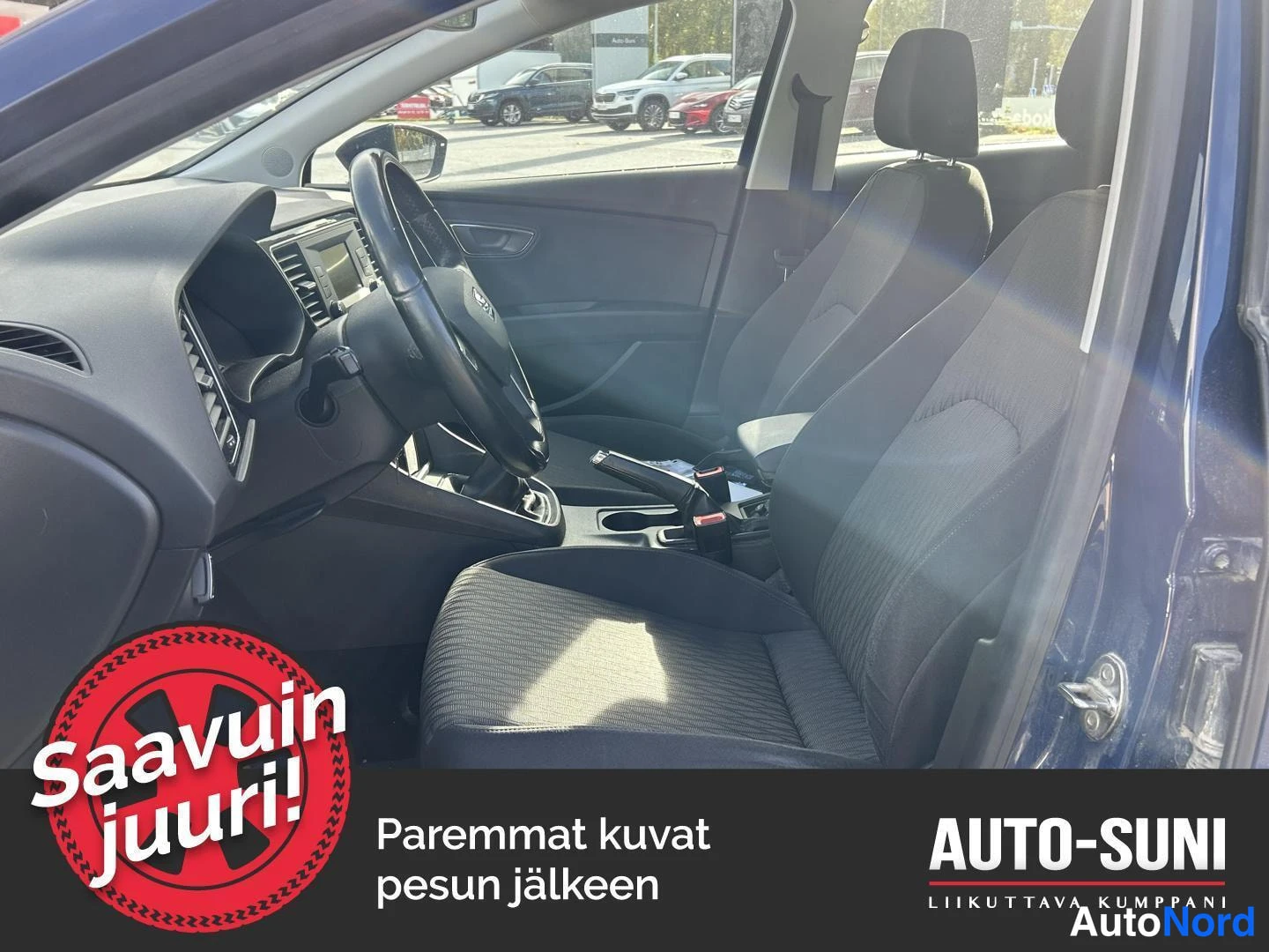 Muu: Seat Leonin etuistuimet ja kojelauta kuvattuna avatusta ovesta; tummaa kankaasta verhoillut penkit, ohjauspyörä ja käsijarru näkyvissä.