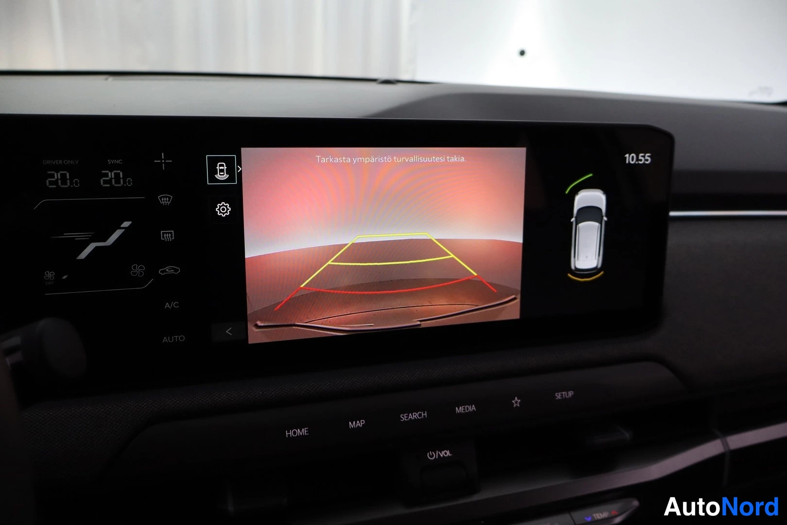 Sivulta: Kuvassa auton infotainment-näyttö, jossa näkyy käänteiskameran kuva apuviivoineen (keltaiset ja punaiset kaaret) ja tekstillä 'Tarkasta ympäristö turvallisuutes