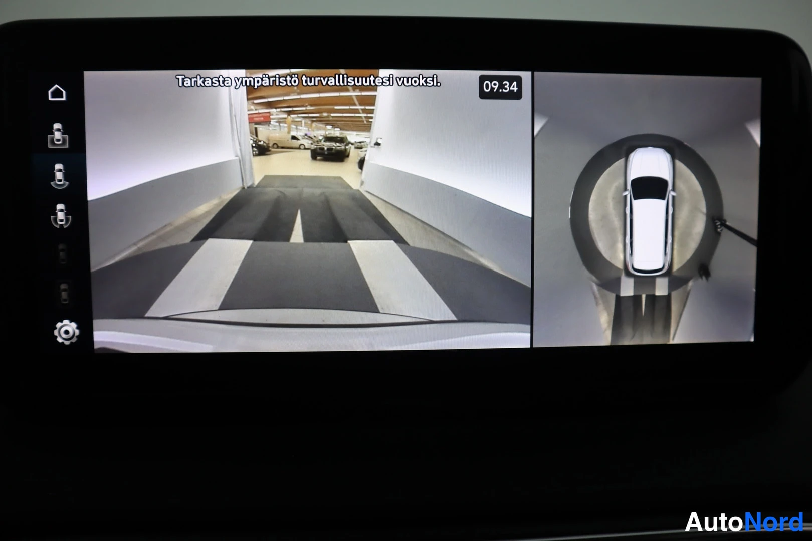 Yksityiskohta: Infotainment-näytöllä näkymä auton ympäriview-kameroista: etukamera ja ylhäältäpäin kuvattu kokonaisnäkymä valkoisesta autosta; teksti 'Tarkasta ympäristö turva