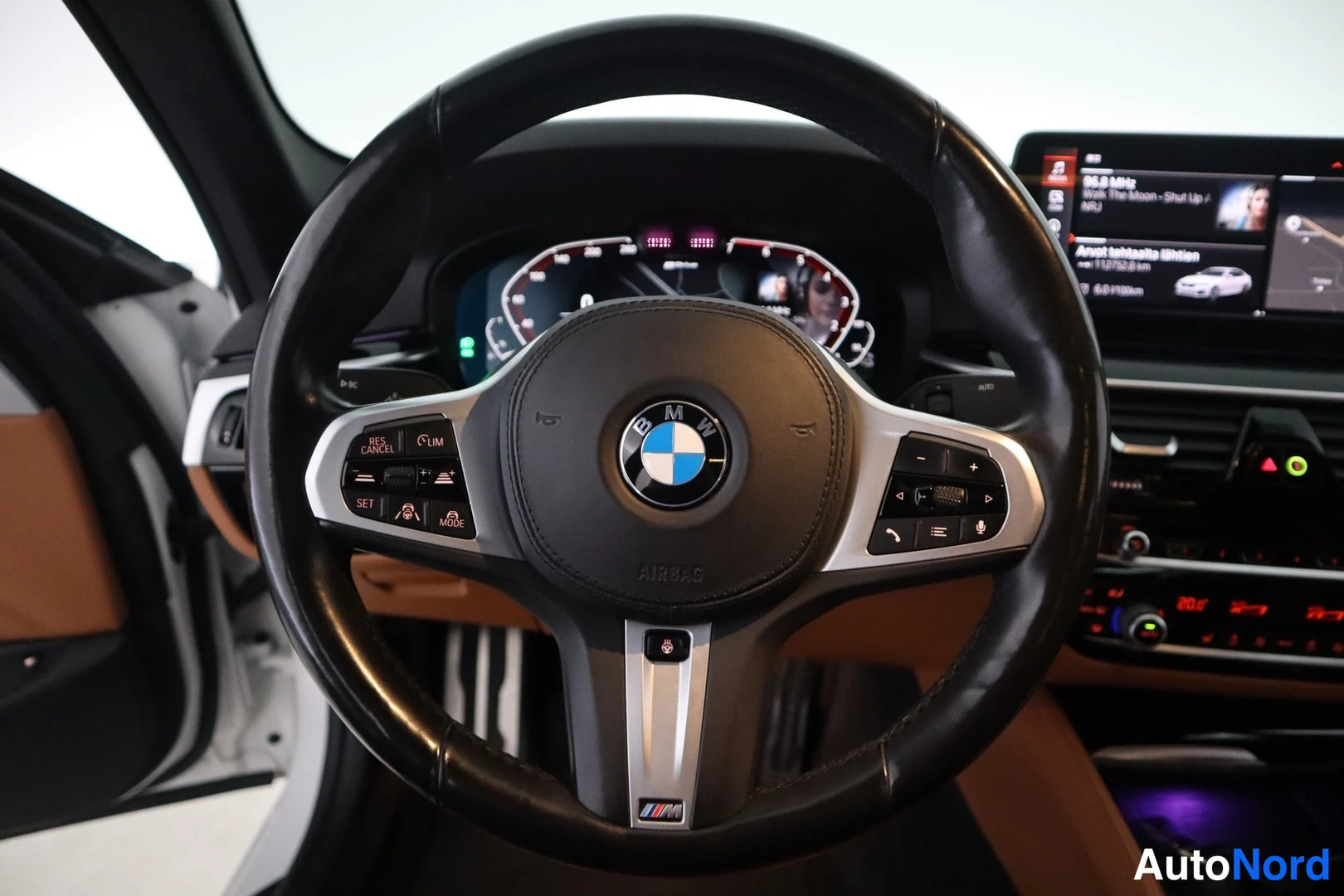 Yksityiskohta: BMW-ohjauspyörä lähikuvassa, digitaalinen mittaristo ja kosketusnäyttö, jolla näkyy radiokanava ja teksti 'Avust ajotietu aktiivinen'。