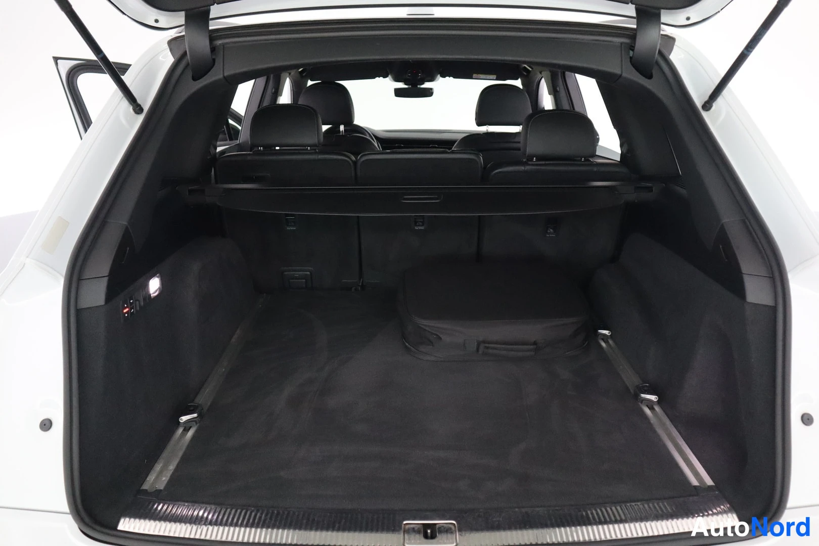 Muu: Audi Q7:n avattu tavaratila: tumma lattiaverho, osittain vedetty tavaratilan kansi ja sähköjohtosarjan pussi lattialla, takapenkin selkänojat taustalla.