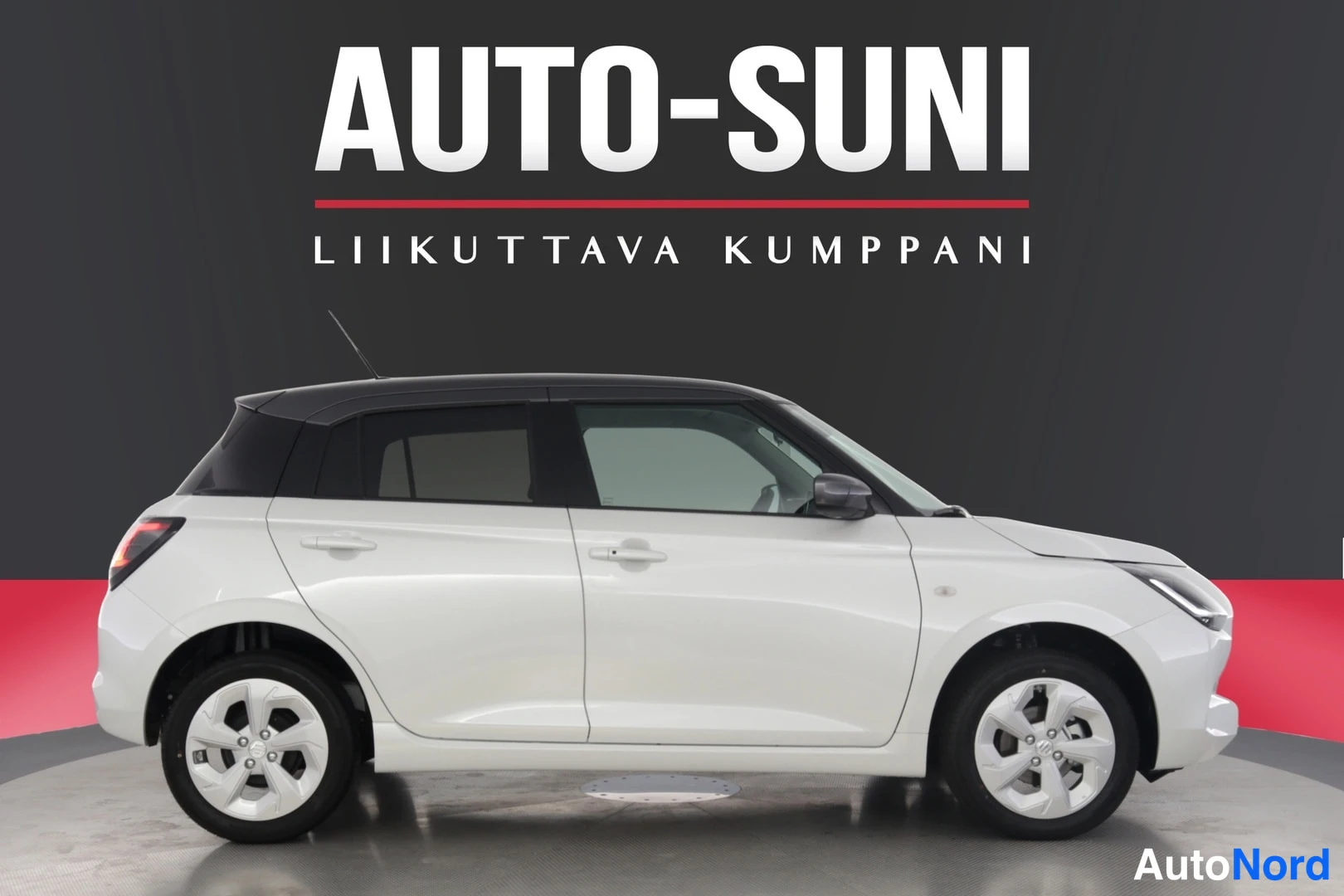 Etuviisto, nokka oikealle: Valkoinen Suzuki Swift sivulta vasemmalta, musta katto, keula näkyy hieman kuvan oikeassa reunassa.