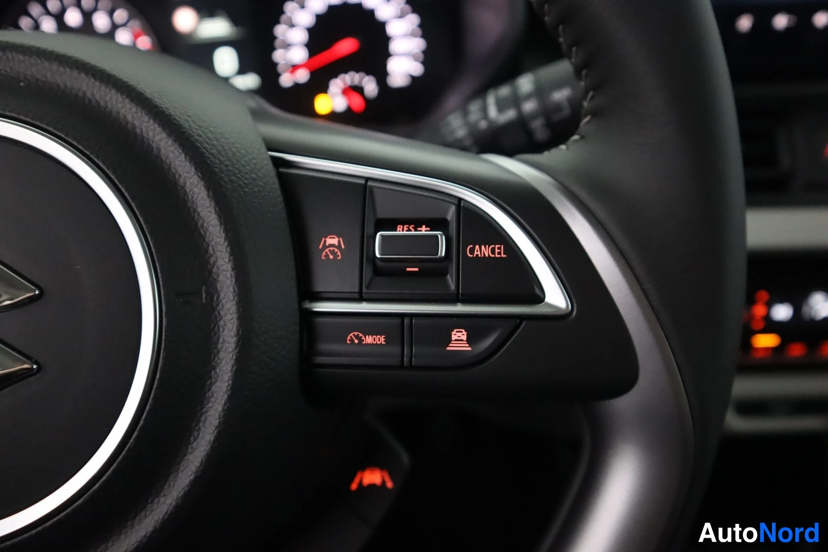Yksityiskohta: Suzuki-merkillisen ratin oikea puoli: cruise control -painikkeet RES+/-, CANCEL, MODE sekä kaistankehotuspainike; taustalla mittaristo valaistuna.