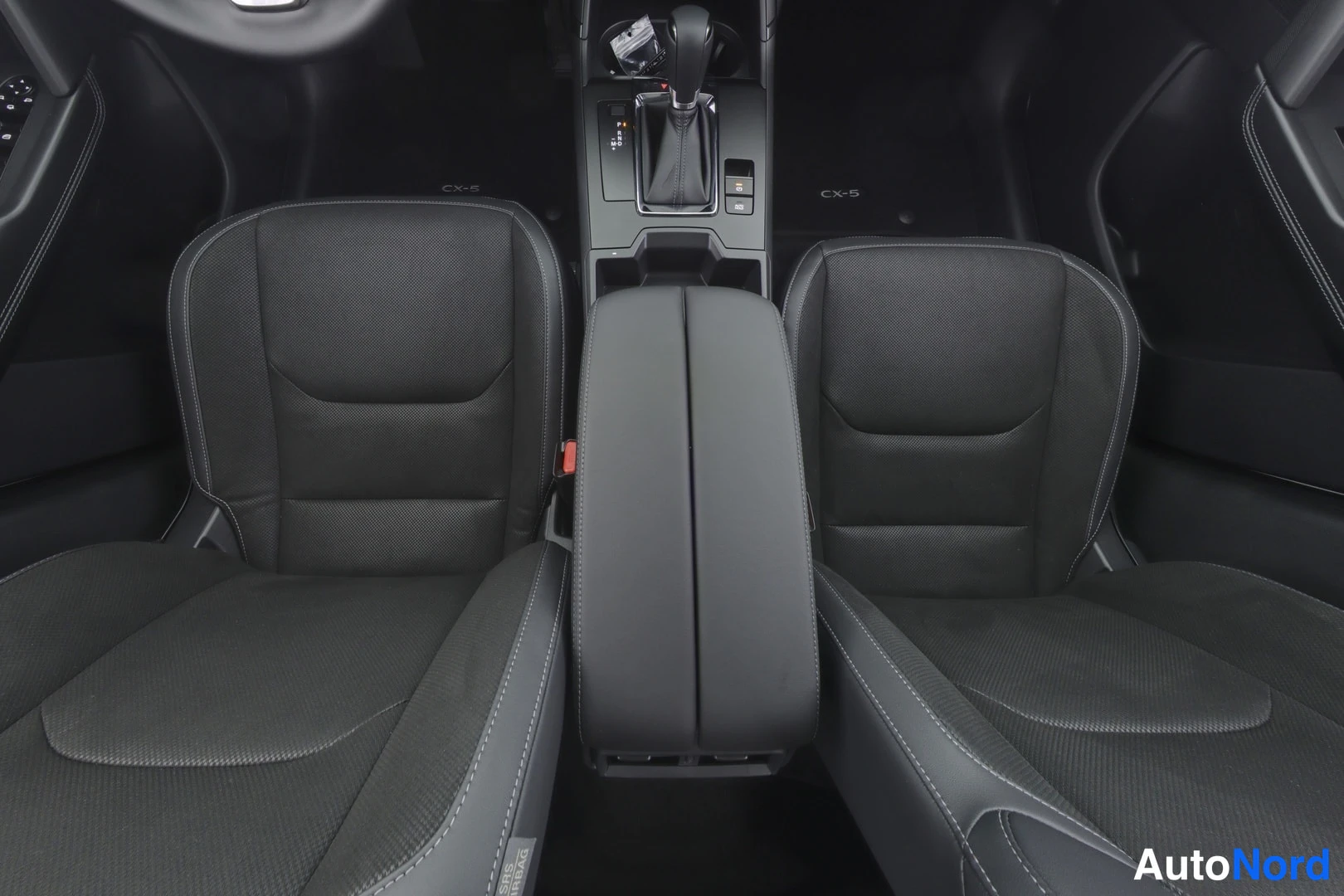 Muu: Mazda CX-5 etupenkit ja keskikonsoli ylhäältä kuvattuna: tummat kankaanpinnaiset penkit, käsituki, automaattivalitsin ja CX-5-matot.
