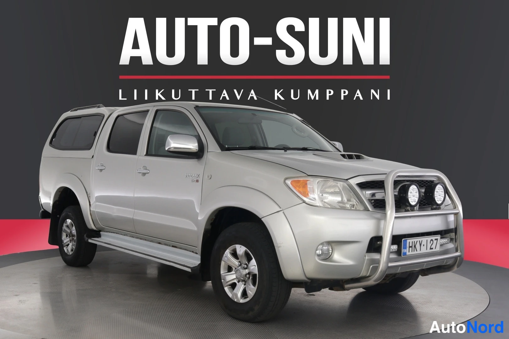 Etuviisto, nokka oikealle: Hopea Toyota Hilux -mallinen avolava-auto kolmannelta etuneljänneskulmalta kuvattuna, etupuskurissa suojakaari lisävaloilla, rekisterikilpi.