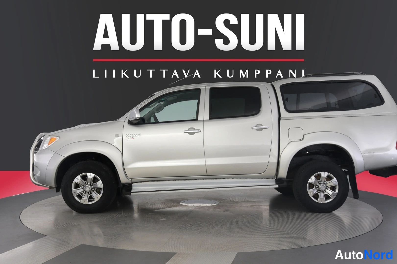 Sivulta: Auto-Suni logo kuvan yläosassa, hopeinen lava-auto sivusta kuvattuna studiovalaistuksessa
