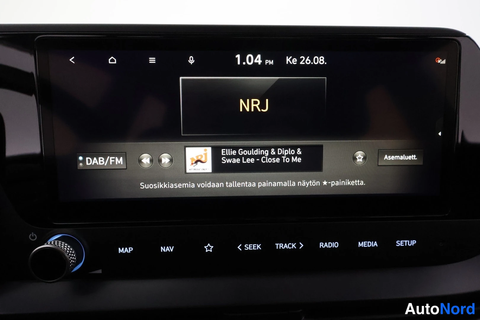 Yksityiskohta: Istumapanelin kosketusnäyttö, jolla toistetaan DAB/FM-radiota asemalta NRJ; näytöllä kello 1.04 PM ke 26.08. ja kappale Ellie Goulding & Diplo & Swae Lee - Clos