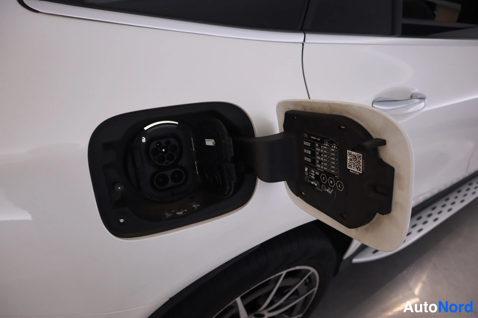 Yksityiskohta: Avattu latausportin luukku auton vasemmassa sivussa; Charge Port -tarra QR-koodeineen näkyvissä.