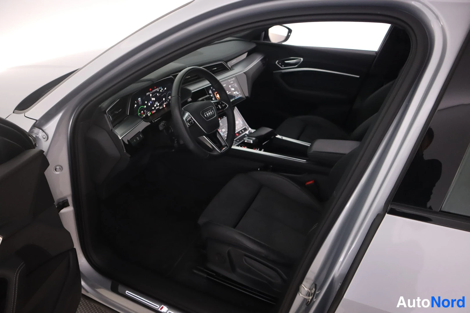 Yksityiskohta: Avatusta etuovesta näkyy hopeisen auton tumma etuistuinosa: Audi-ohjauspyörä, digitaalinen mittaristo, keskikonsolin näytöt ja tummat penkit.