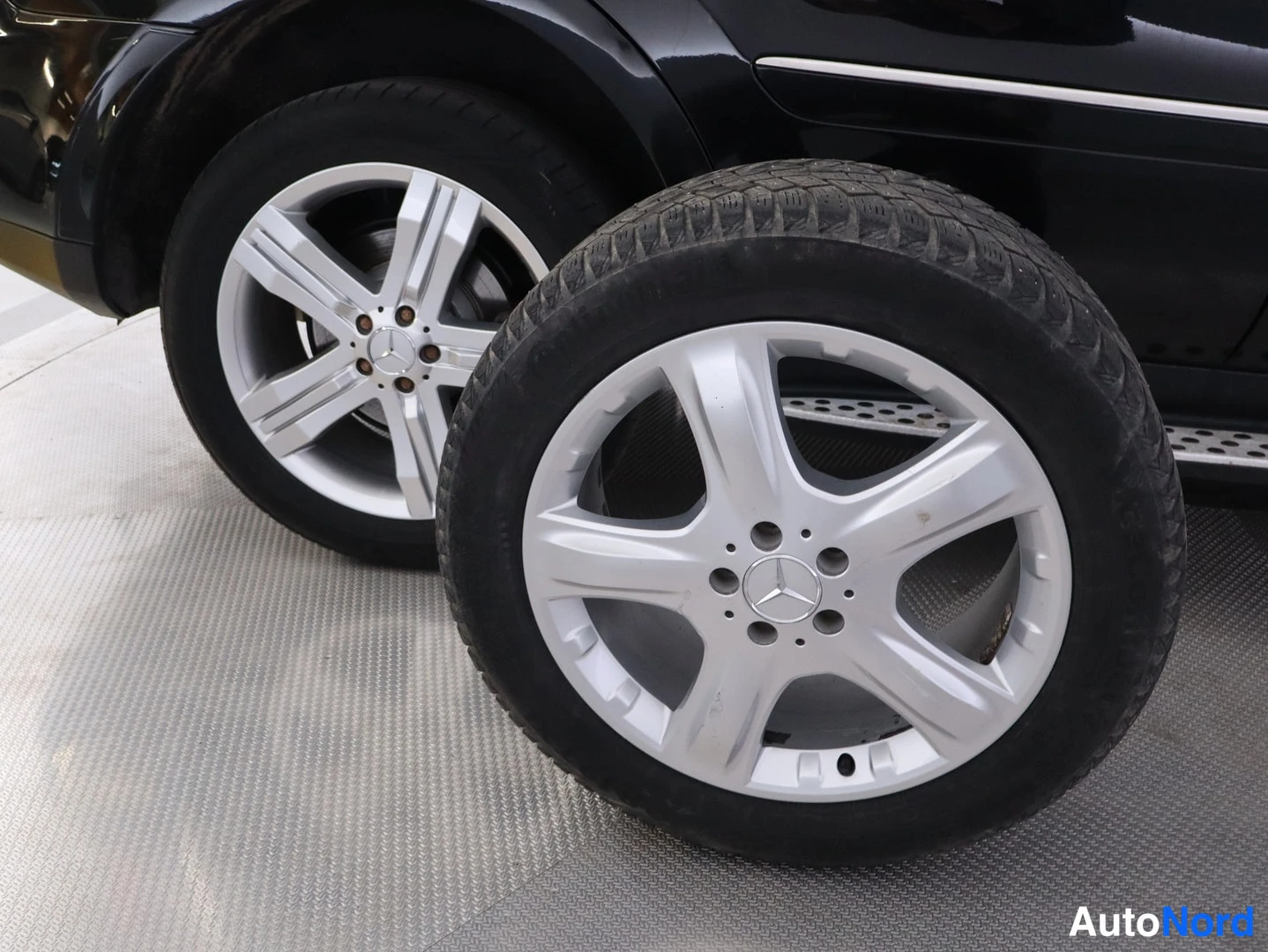 Yksityiskohta: Renkaiden ja hopeisten Mercedes-merkitysten kevytmetallivanteiden lähikuva mustan auton vieressä.