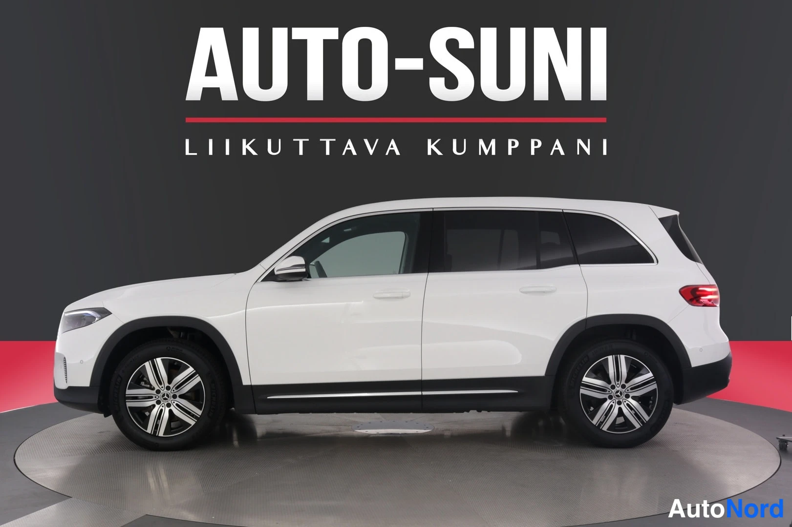 Etuviisto, nokka vasemmalle: Mercedes-Benz EQB -farmari-tyyppinen sähköauto valkoisena kulmassa kuvattuna autosalissa, kylki ja etunosa näkyvissä