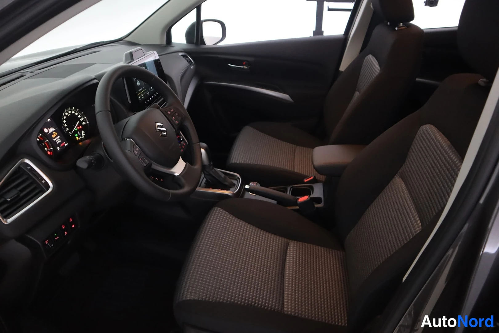 Yksityiskohta: Sisätilakuva etuistuimilta: Suzuki steering wheel, black dashboard, infotainment display, fabric front seats in dark grey, automatic gear lever visible.
