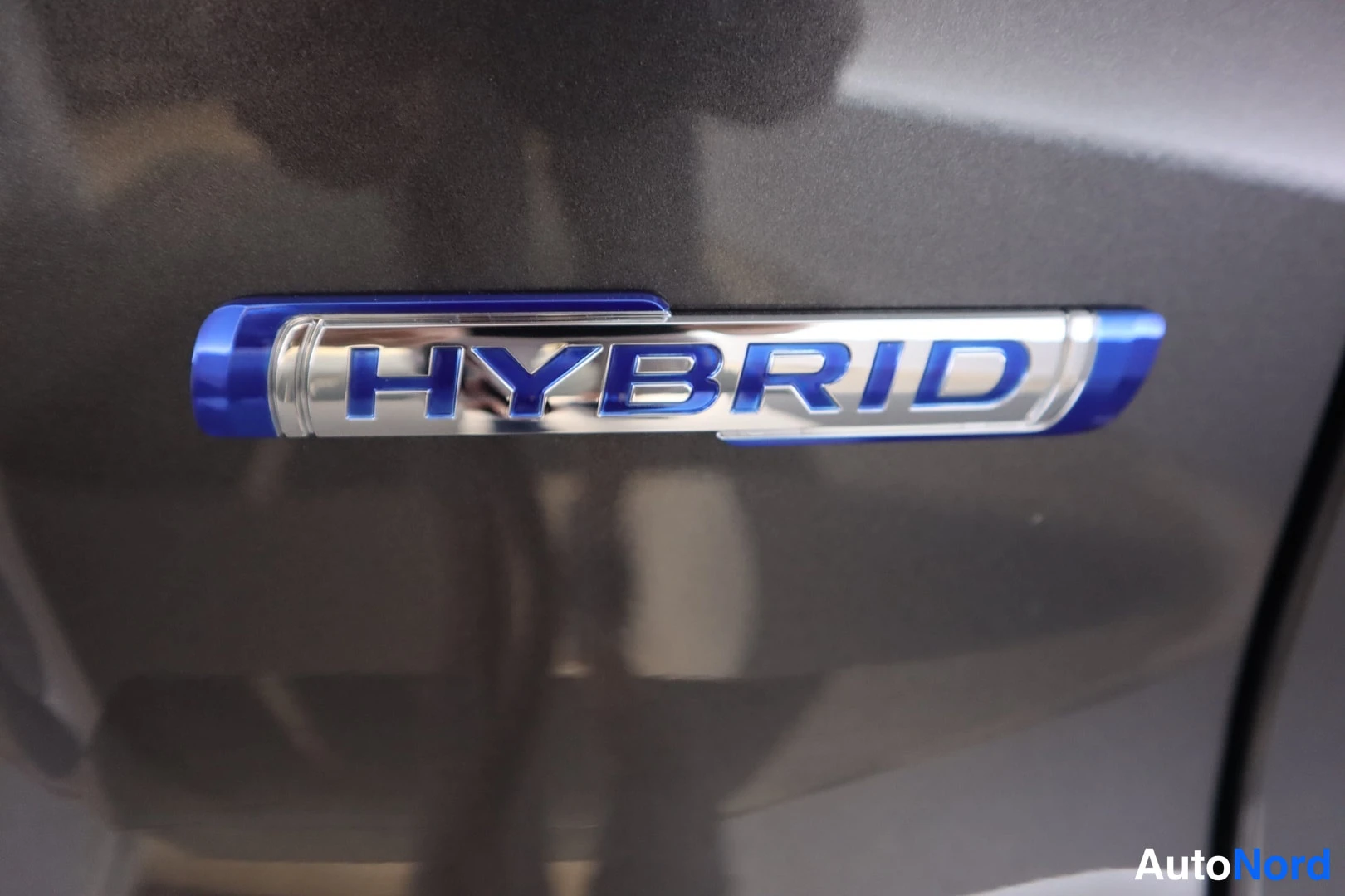 Yksityiskohta: Sininen HYBRID-embleemi auton tummassa koripaneelissa.